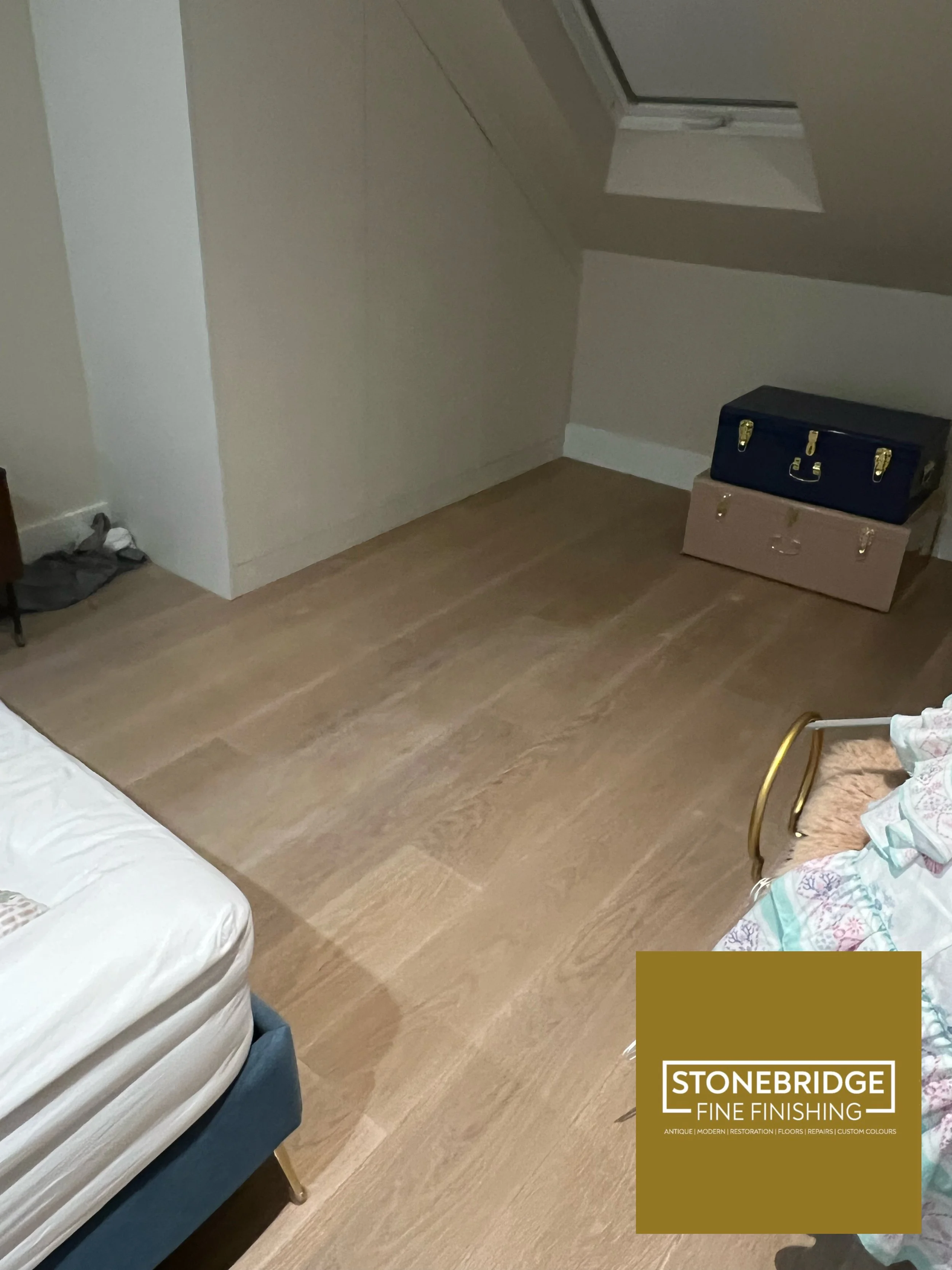 Room with wooden flooring, a bed with a white mattress, a chair, a stack of two suitcases, and a logo in the bottom right corner for Stonebridge Fine Finishing.