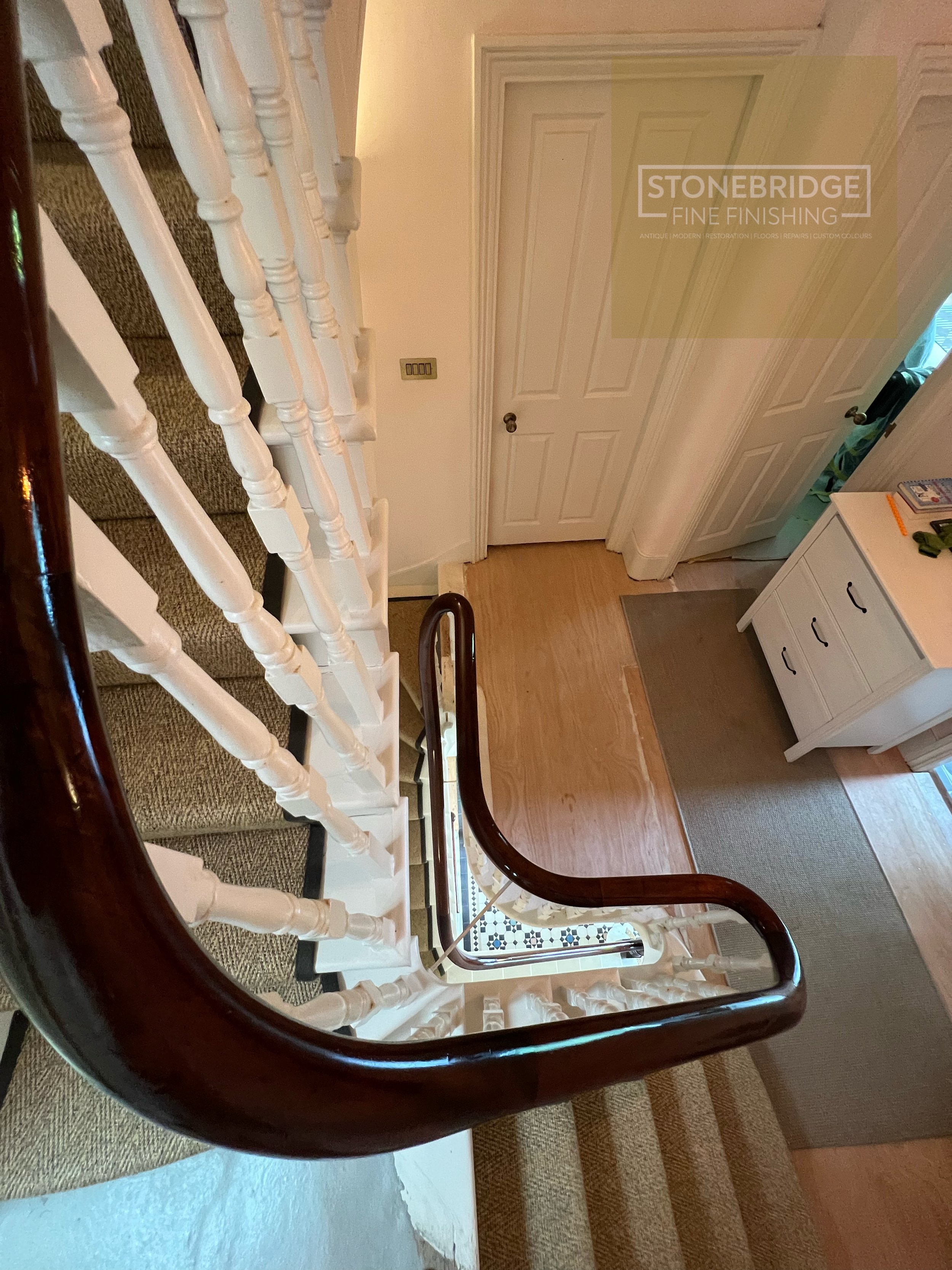 View of a staircase with white balusters and a dark wooden handrail, looking down towards the entryway with a closed door and a dresser nearby.