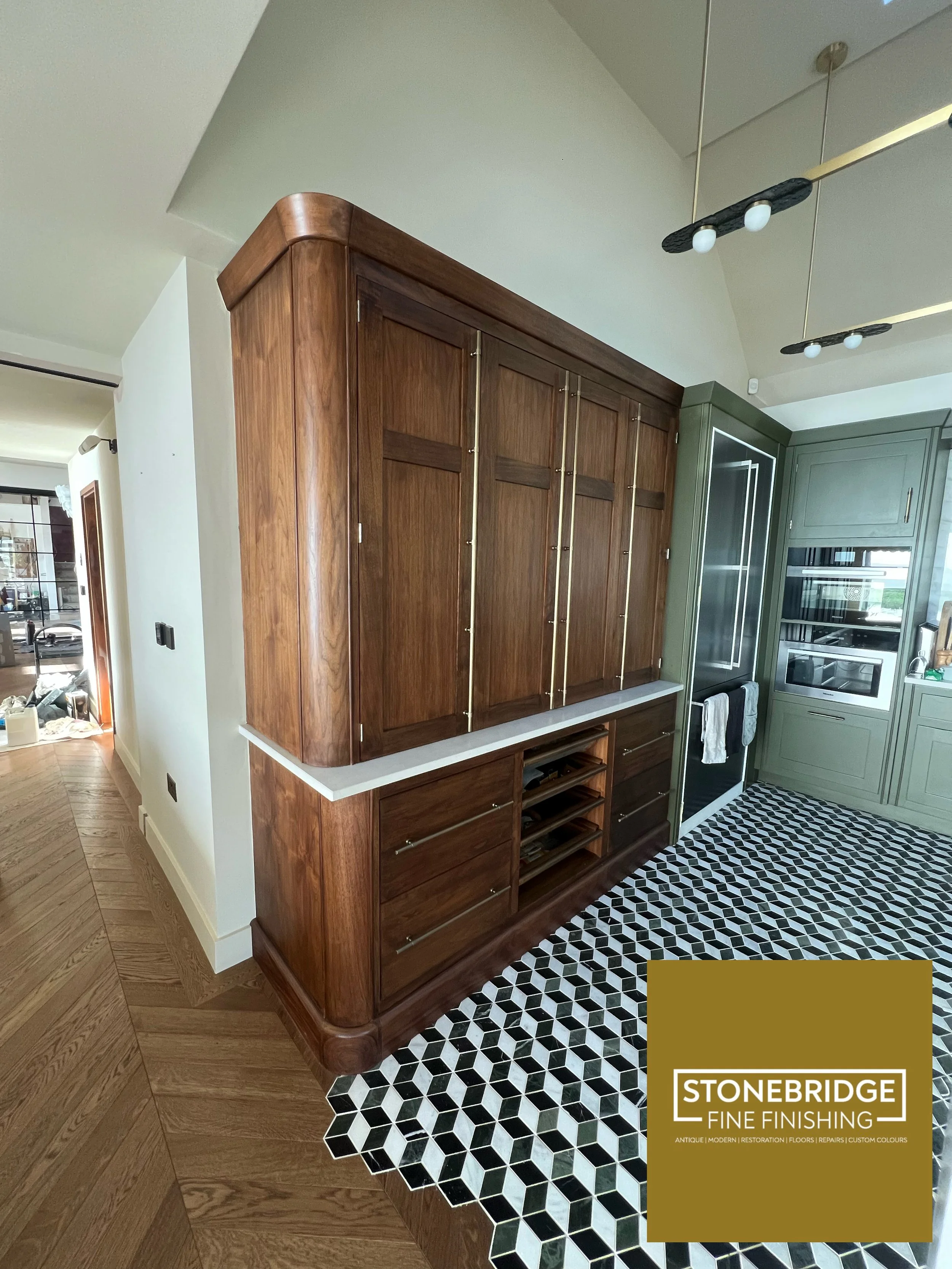 Interior of a home showing a wooden built-in cabinet with brass accents, a section of a kitchen with green cabinetry and a black and white patterned tile floor, and a modern black and brass ceiling light fixture.