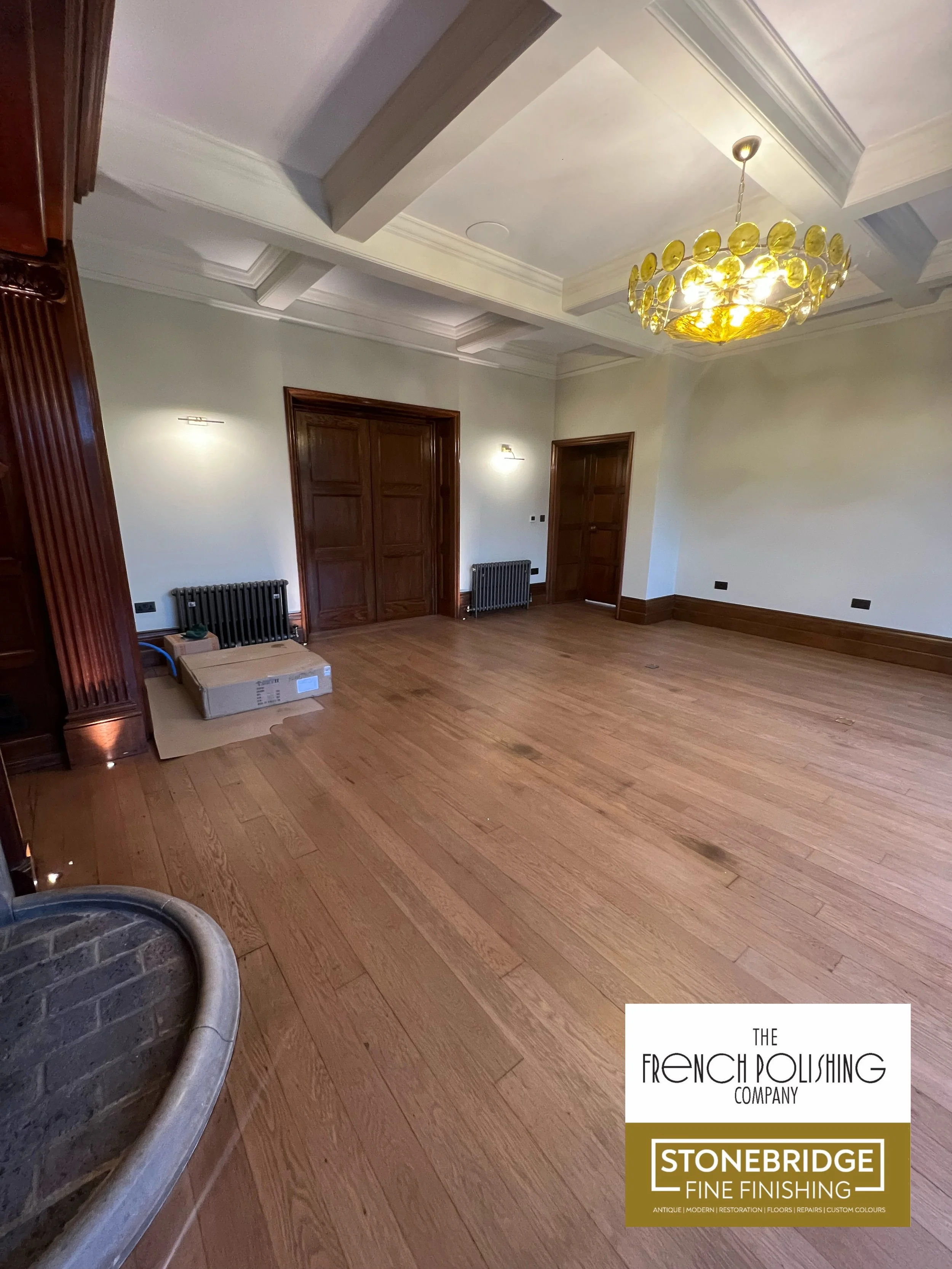A room with wooden floors and wooden doors. There is a chandelier hanging from the ceiling and some radiators on the walls. In the bottom right corner, there is a logo for The French Polishing Company, Stonebridge Fine Finishing.