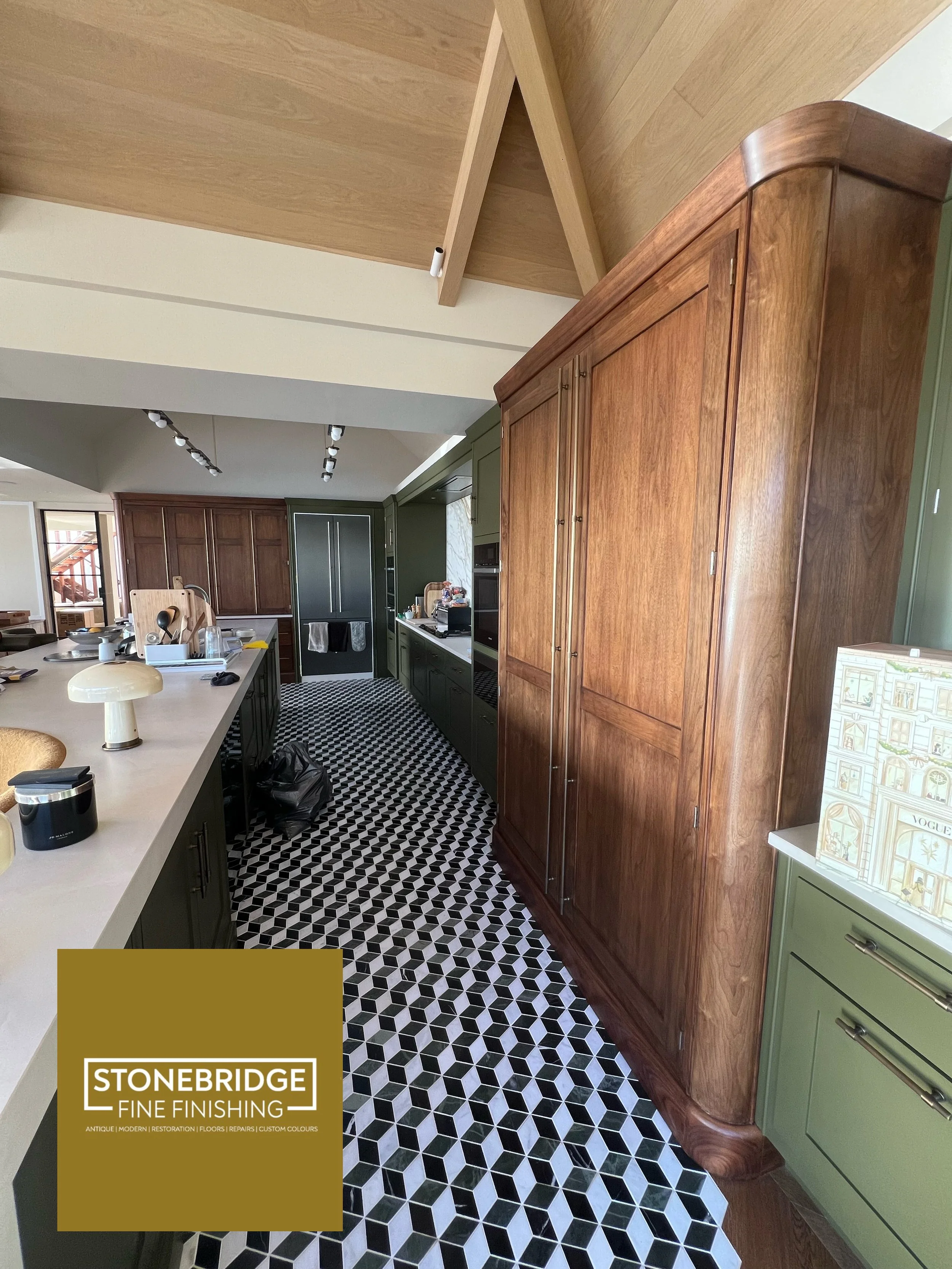 A modern kitchen with a black and white geometric patterned tile floor, oak and green cabinetry, a large wooden pantry, and a kitchen island with various items on it.