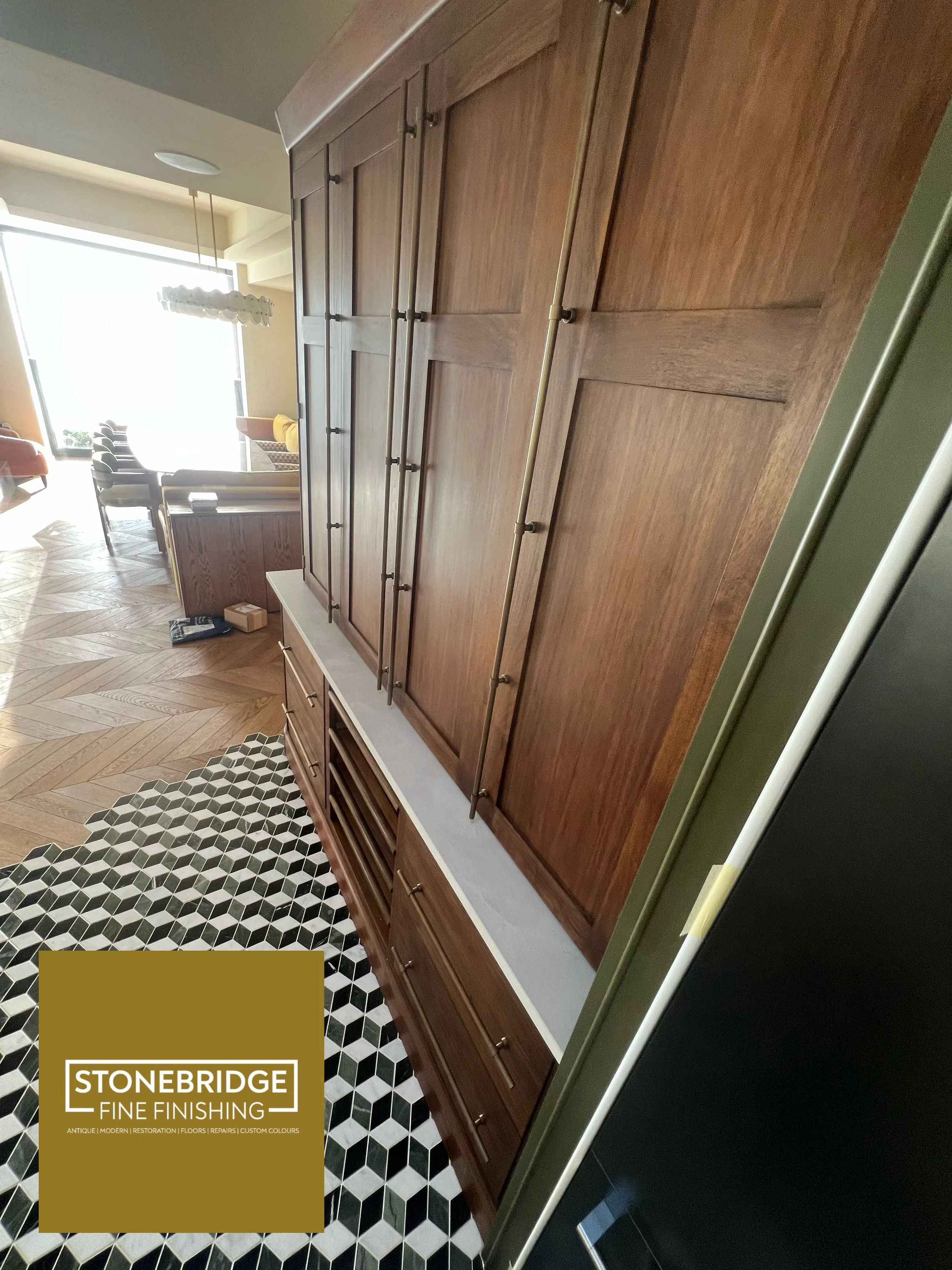A large wooden cabinet with metal handles in a modern kitchen, with a black and white hexagonal tile floor and a living room area with chairs and a large window in the background.