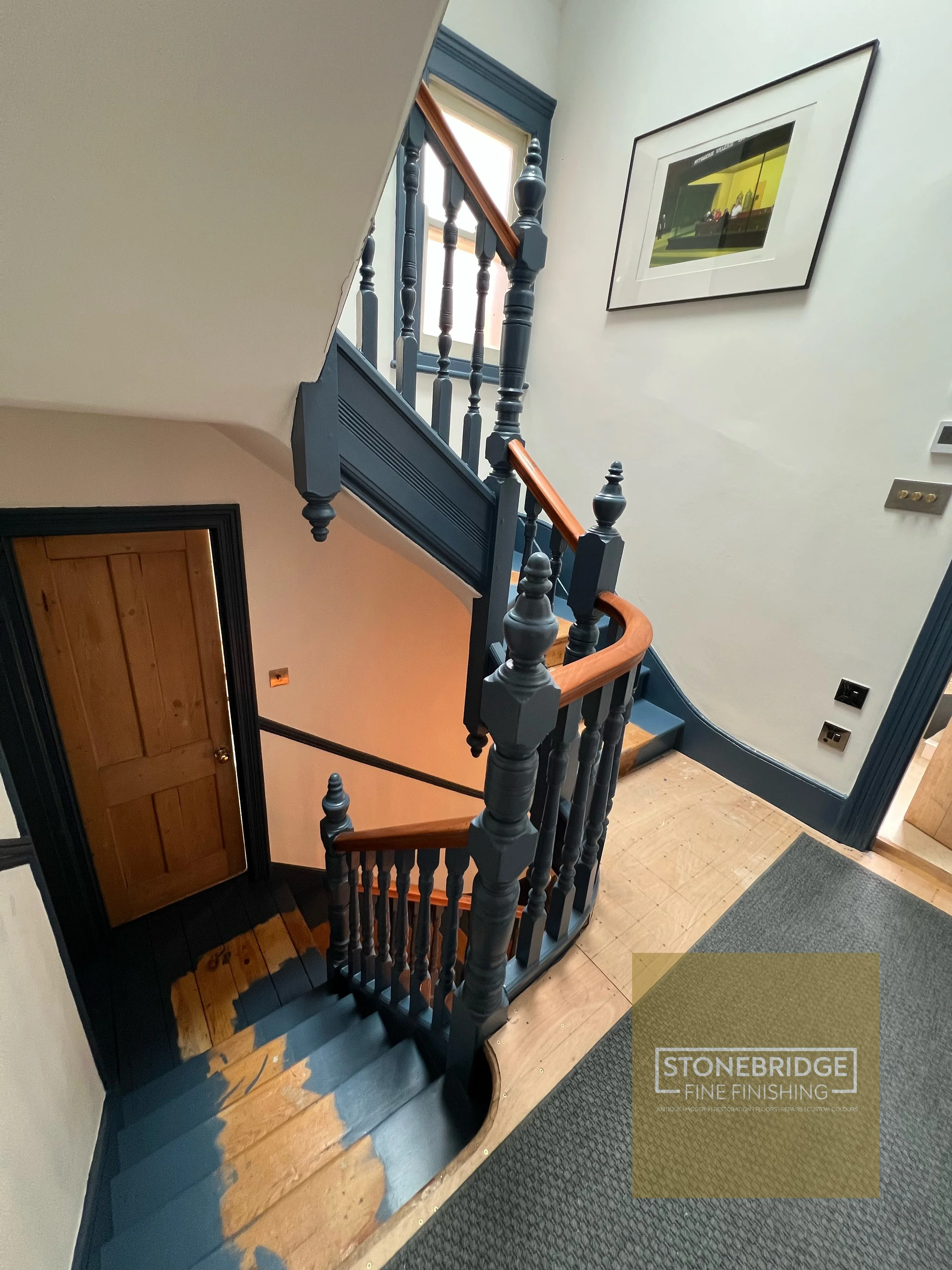 Interior staircase with painted black balustrades, wooden handrail, and partially finished wooden stairs. A framed picture hangs on the wall, and a door with wooden panels is visible below the staircase. The floor has wood and carpeted sections, with