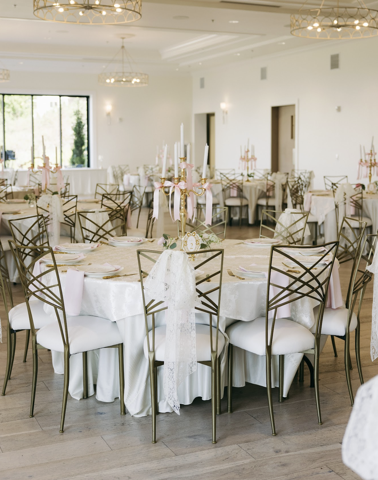 Elegant banquet hall with round table settings, decorated with white tablecloths, pink ribbons, gold flatware, fine china, and a tall gold candelabrum centerpiece with pink ribbons and white candles. Multiple tables arranged throughout the room with matching chairs, large windows in the background, and modern chandeliers overhead.