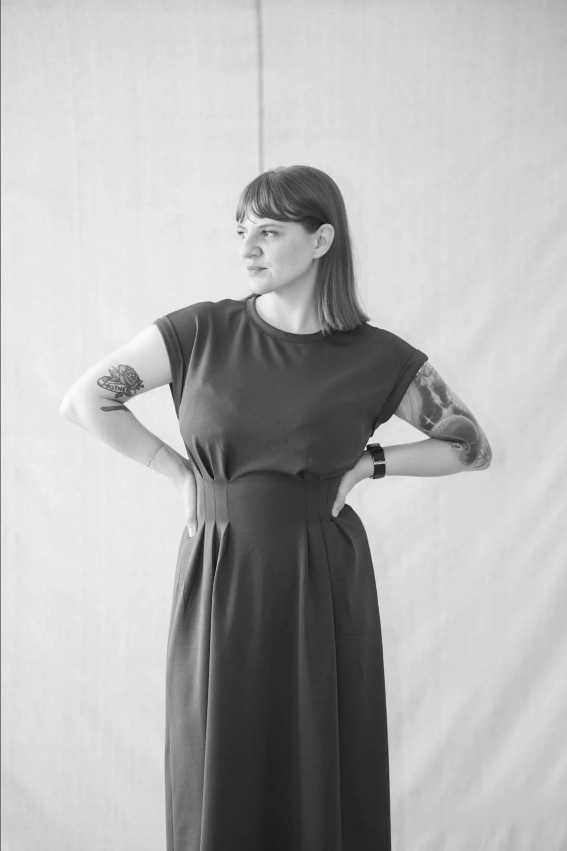 A woman with short dark hair and tattoos on both arms, wearing a black dress, stands with her hands on her hips, looking to her left against a plain wall.