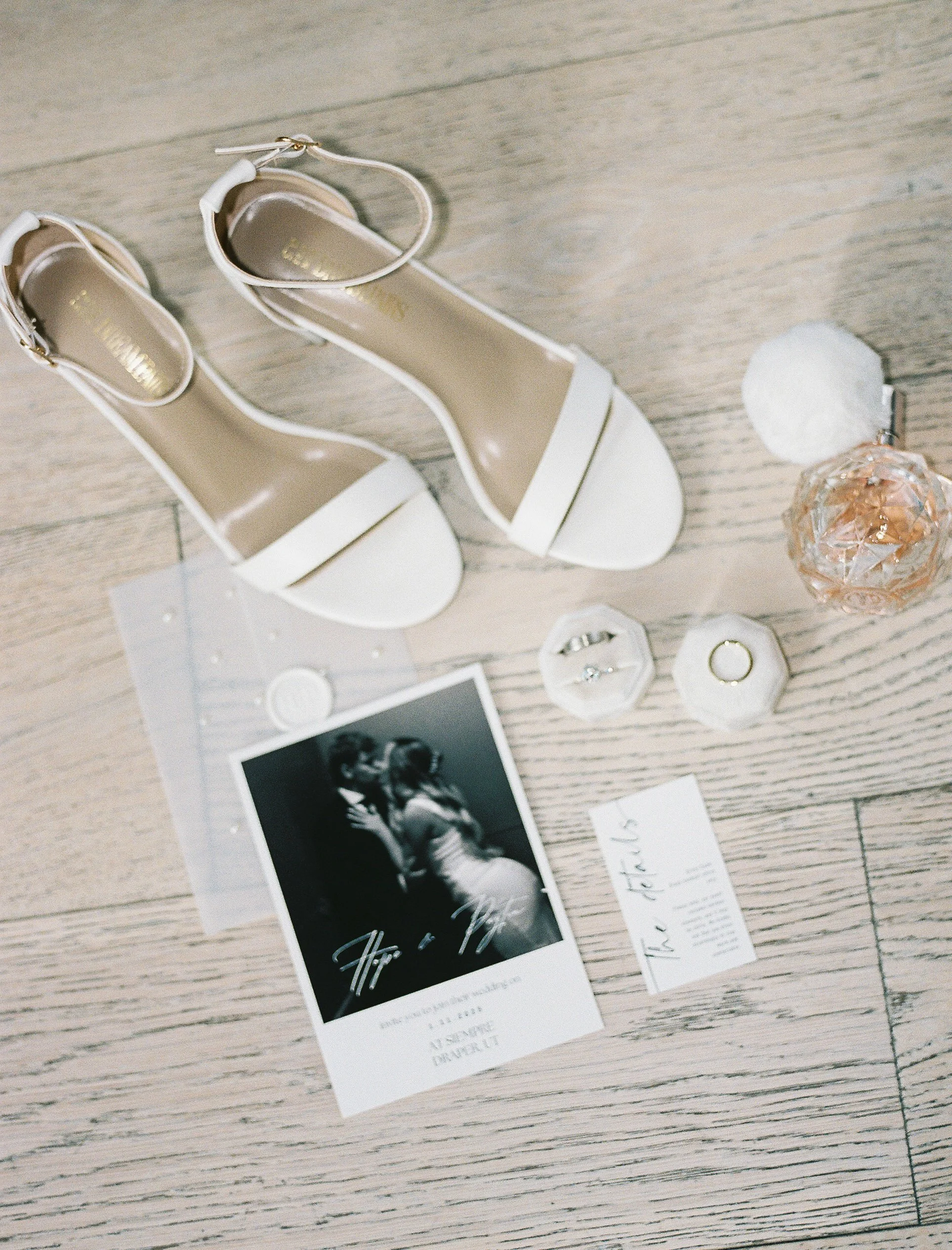 A pair of white high-heeled shoes with ankle straps on a wooden floor, surrounded by black-and-white photo of a couple, jewelry, a perfume bottle, and a handwritten note.