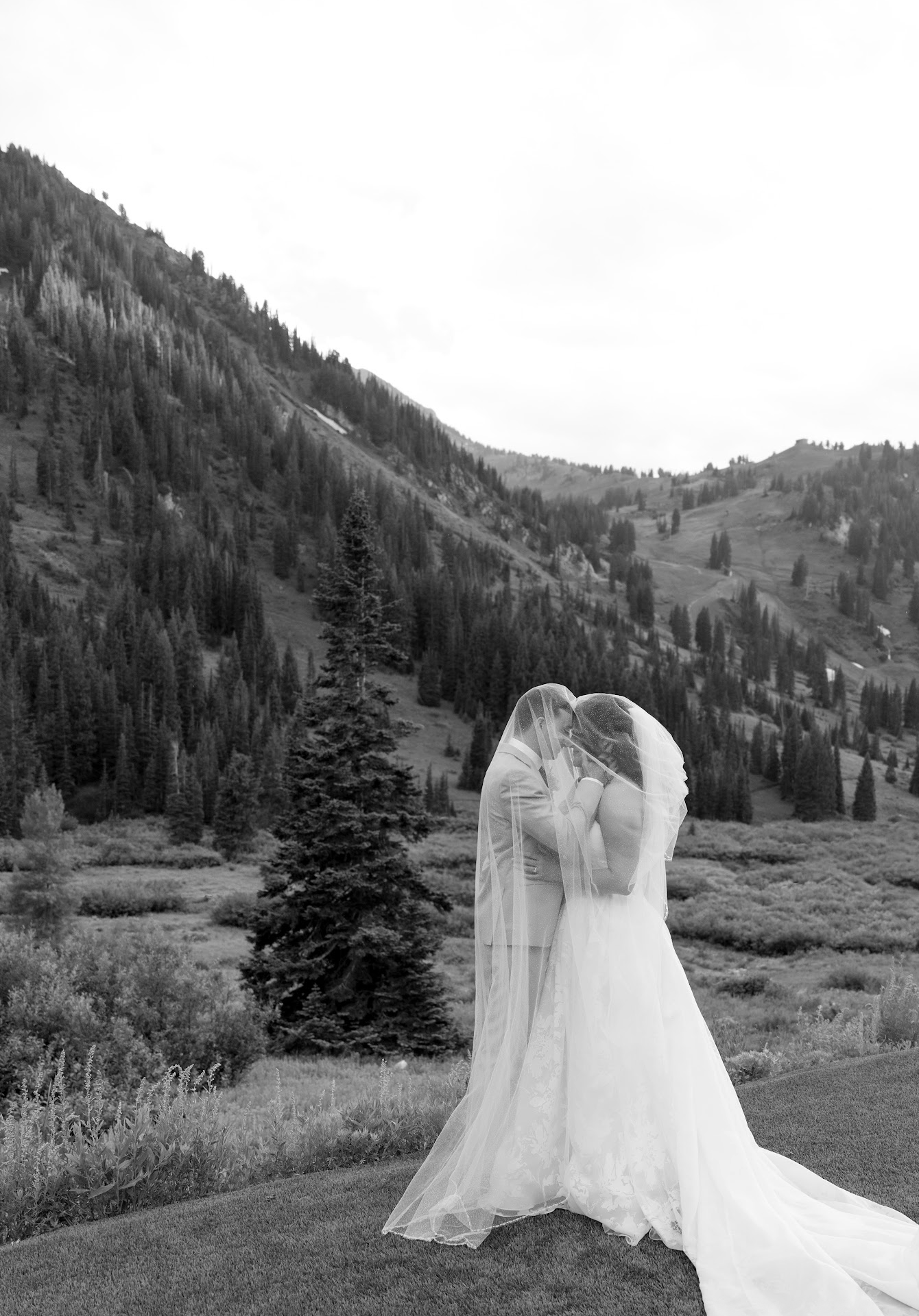 A bride and groom dressed in wedding attire, embracing and kissing outdoors with a mountainous landscape and pine trees in the background.