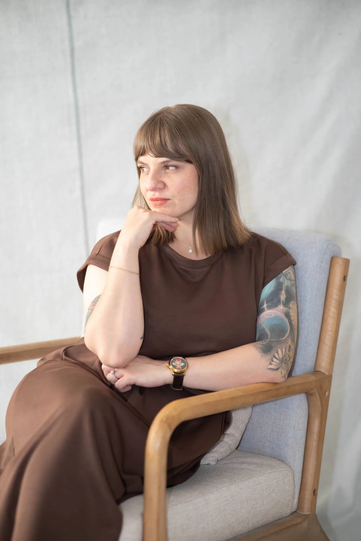 A woman with brown hair and tattoos on her left arm, sitting on a beige armchair against a plain wall, resting her chin on her hand, looking thoughtfully to the side, wearing a brown outfit and a watch.