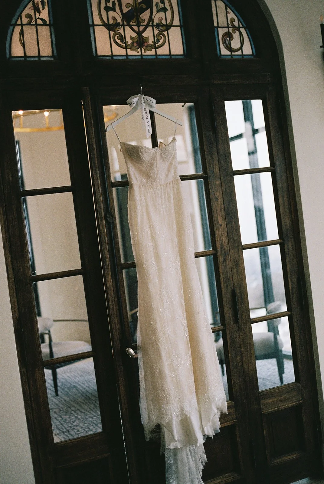 A white wedding dress hanging on a white hanger with a ribbon, displayed in front of a wooden door with glass panels and decorative stained glass at the top.