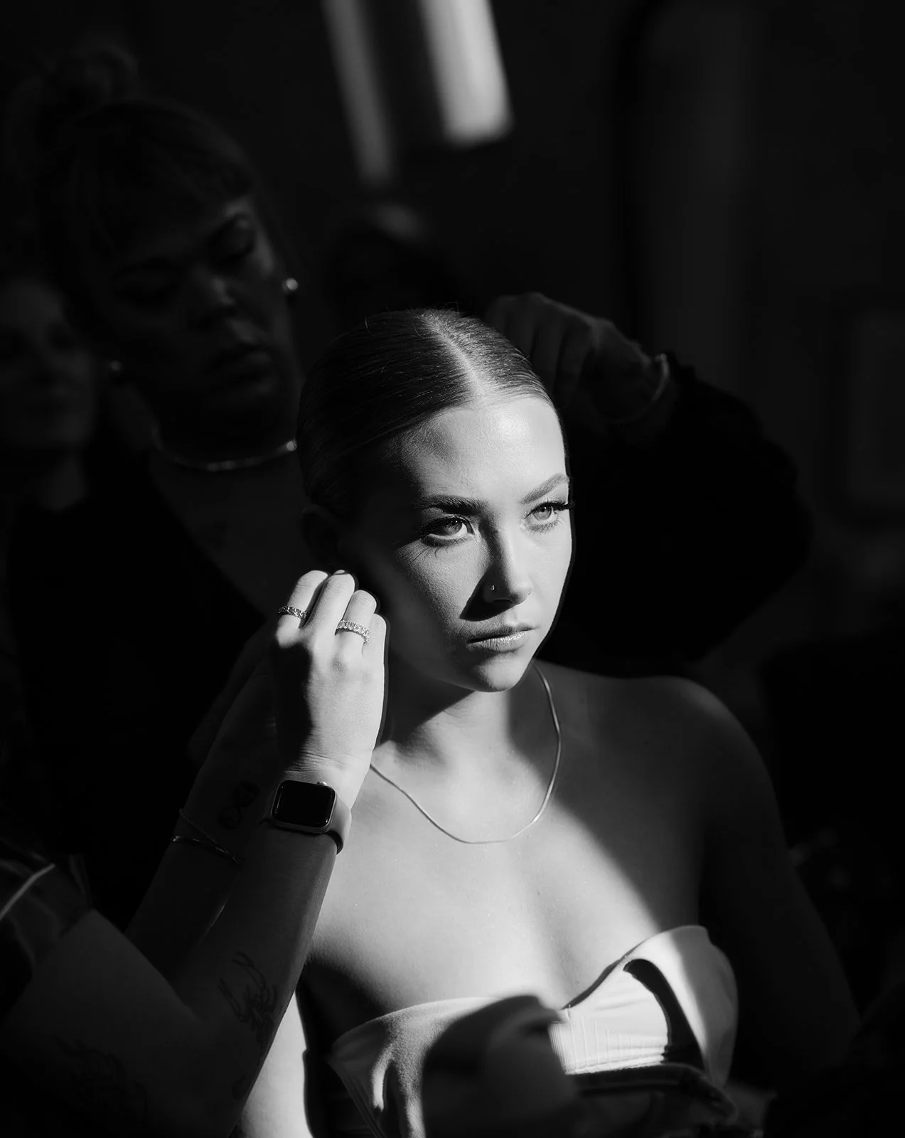 A woman with dark hair styled back, sitting in a well-lit spot, being attended to by a stylist or makeup artist. The stylist's hand adjusts jewelry on the woman's ear, and the woman gazes thoughtfully into the distance with a serious expression. The scene is in black and white.