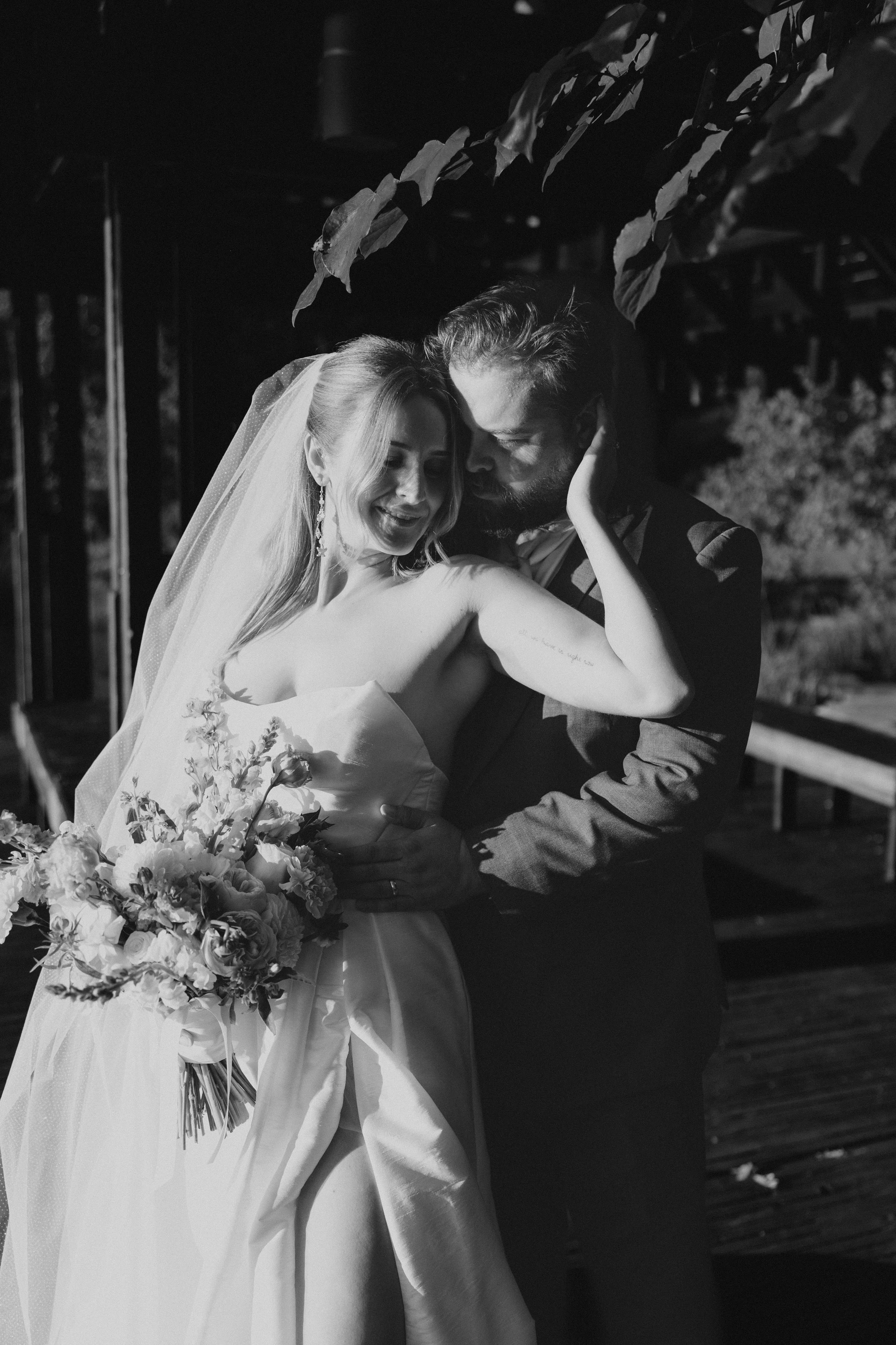A black and white photograph of a bride and groom in a romantic moment, with the bride holding a bouquet of flowers and wearing a wedding dress and veil, while the groom is dressed in a suit. They are close together, with the groom touching the bride's face and forehead in an intimate pose, under some leaves or foliage.