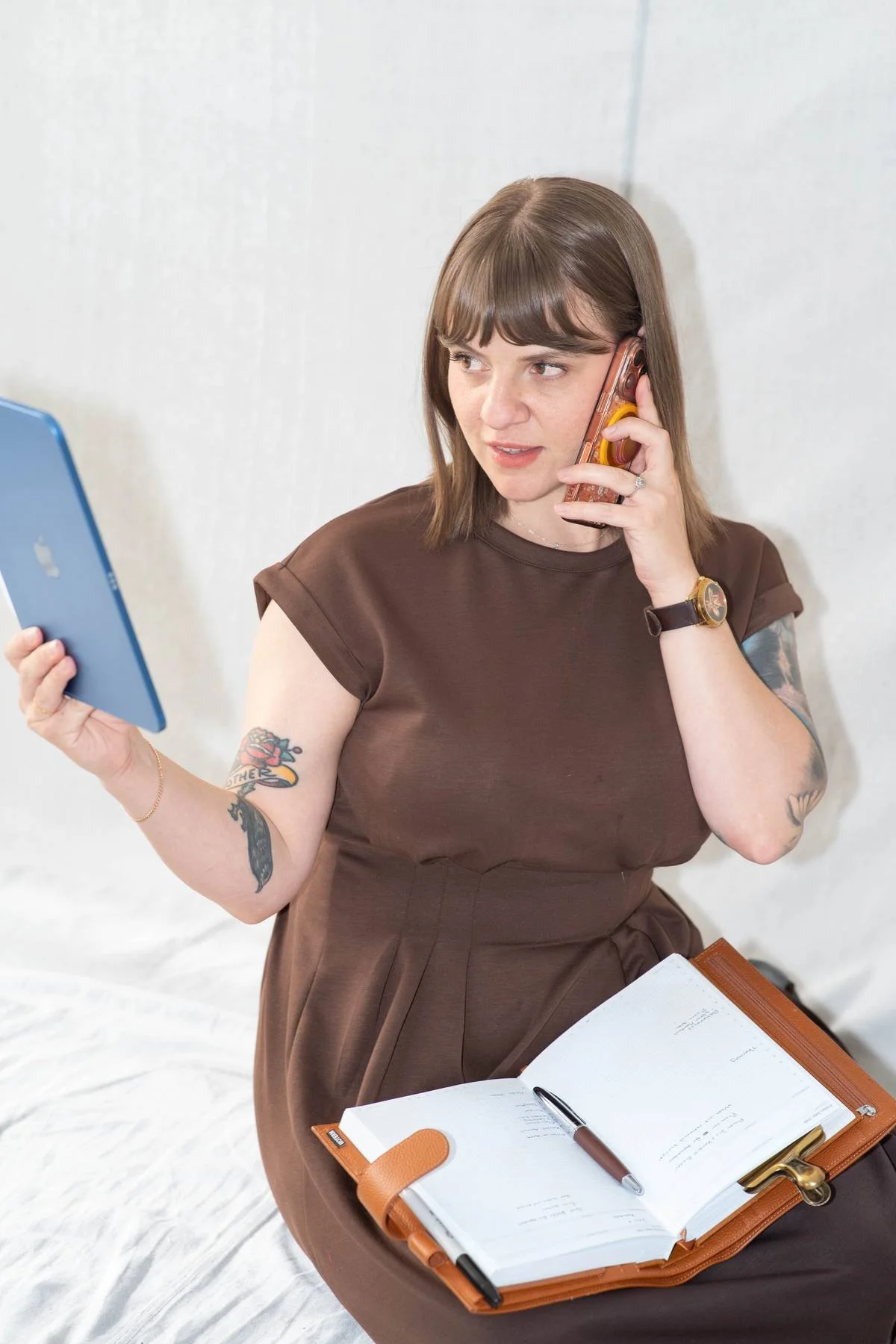 Woman in brown dress talking on a phone while holding a blue tablet, with an open planner and pen on her lap, seated against a white wall.