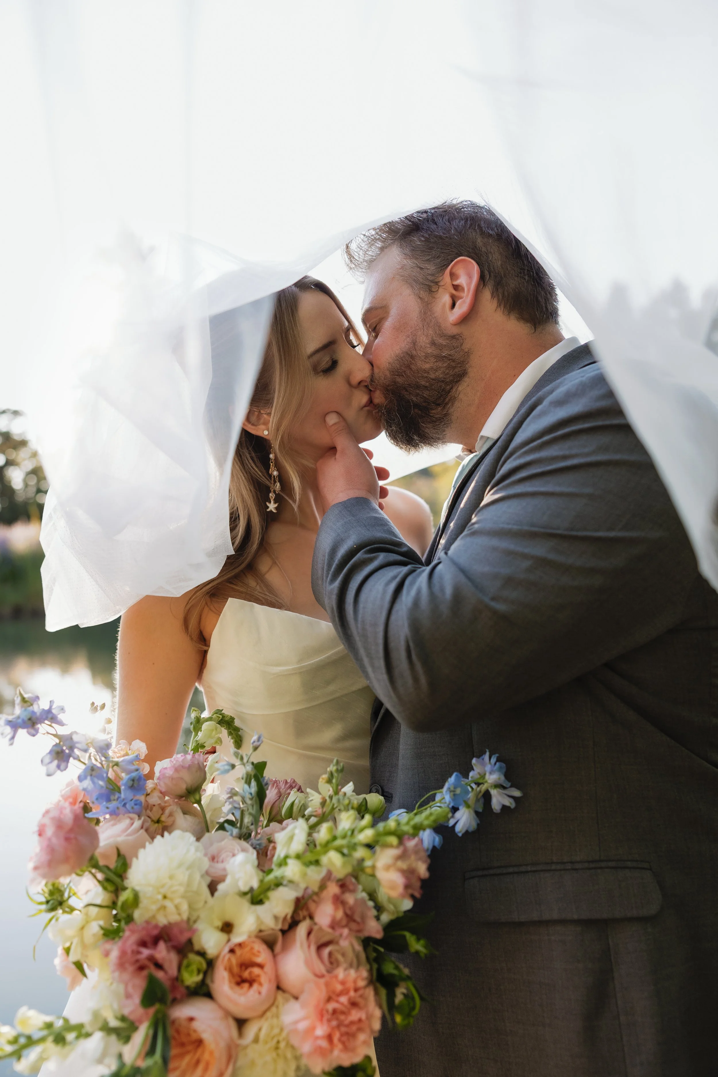 A man and woman kissing during a wedding ceremony outdoors, the woman wearing a white wedding dress and veil, holding a large bouquet of pink, white, and purple flowers, with a blurred natural background.