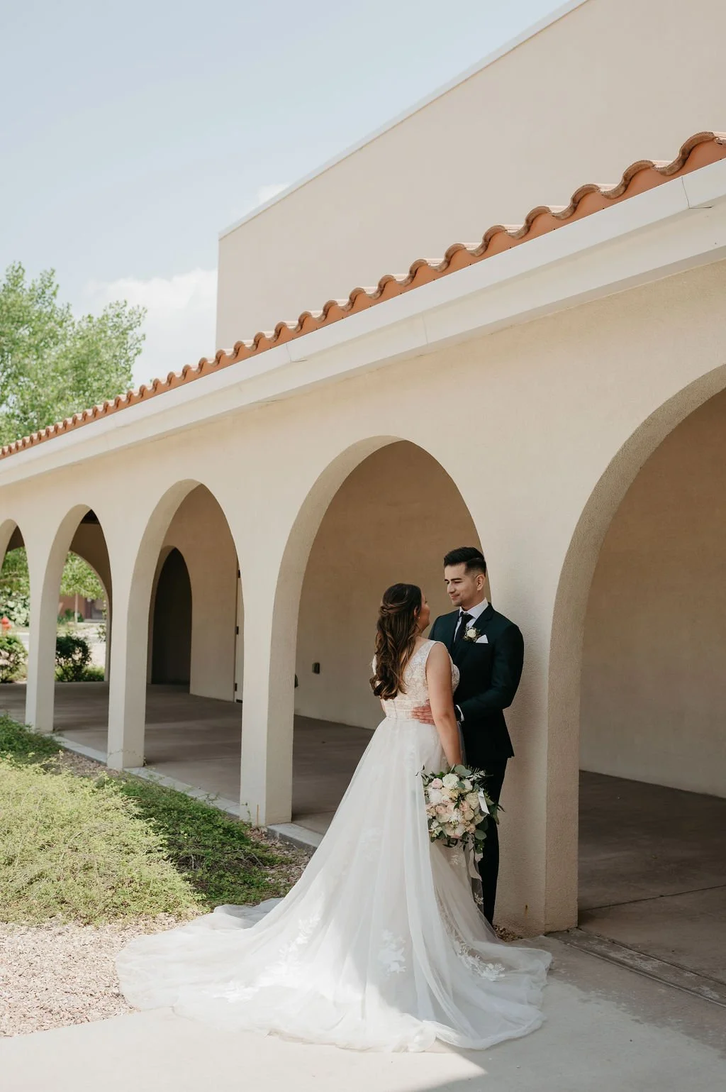 A bride and groom share a romantic moment outdoors under an arched walkway, with the bride holding a bouquet of flowers.