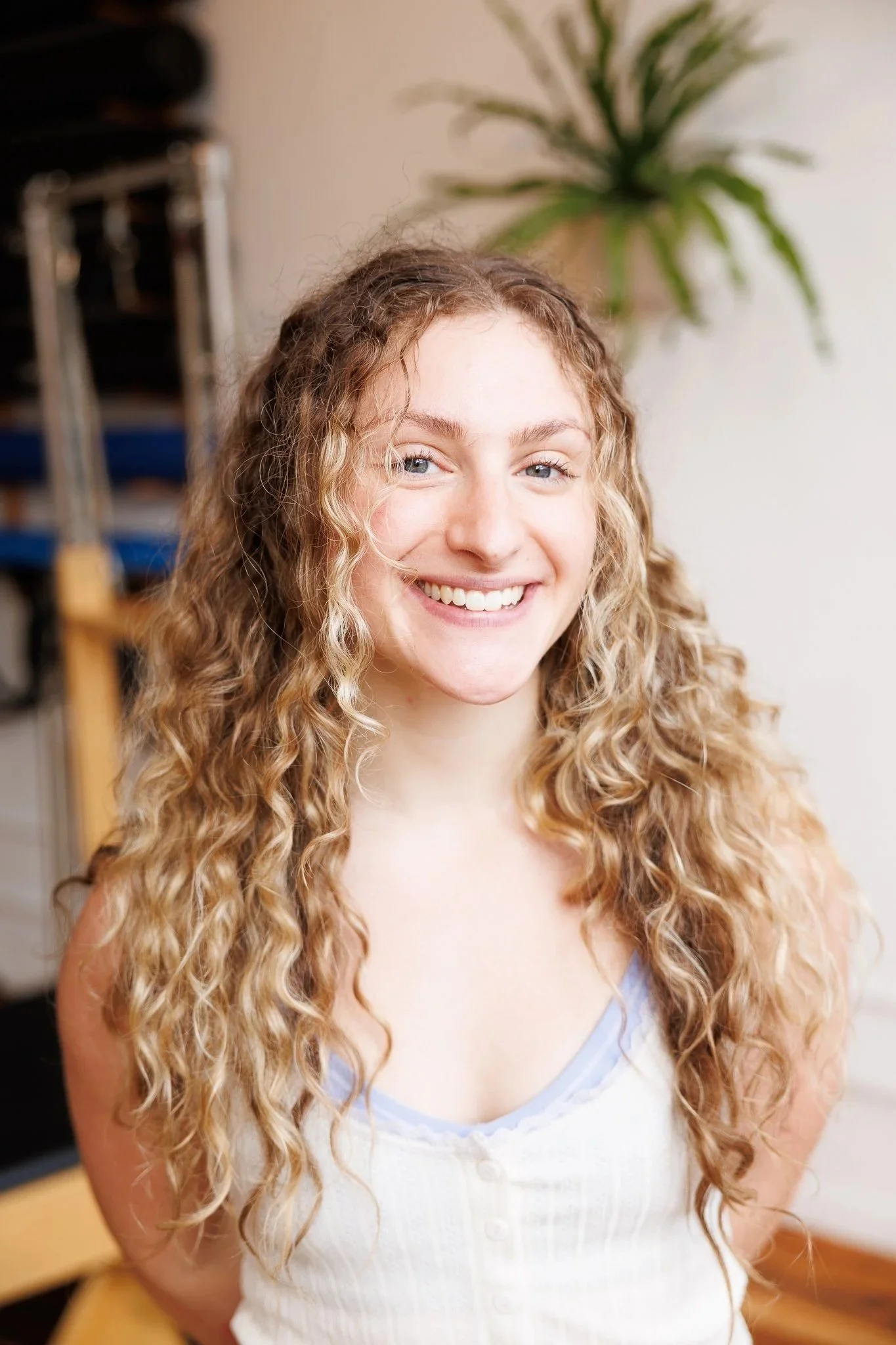 A young woman with long, curly blonde hair smiling and sitting in a fitness or yoga studio with black mats and a wooden floor.
