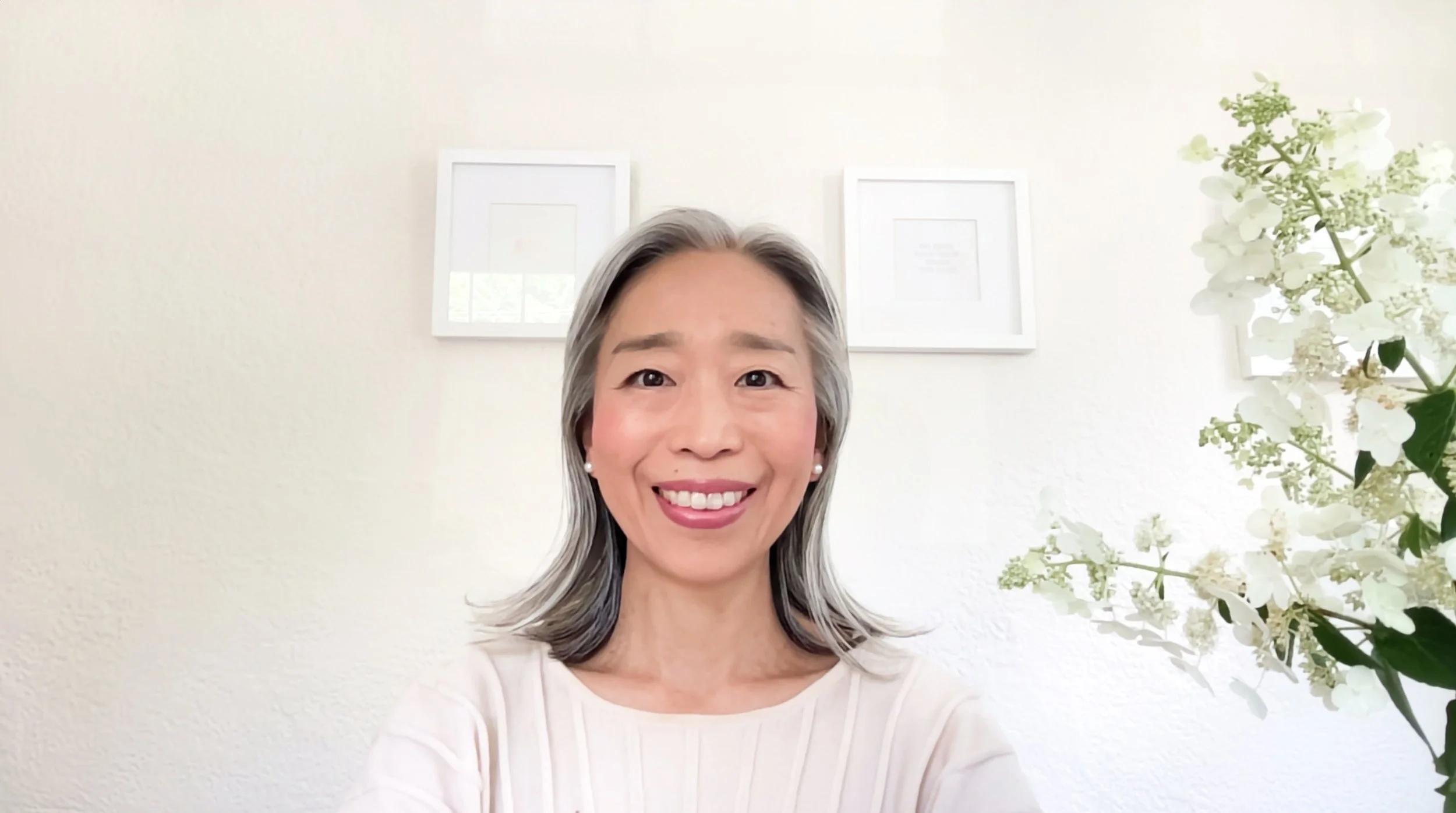 Smiling woman with shoulder-length gray hair taking a selfie in a bright room with white walls and framed pictures behind her, and white flowers on her right.