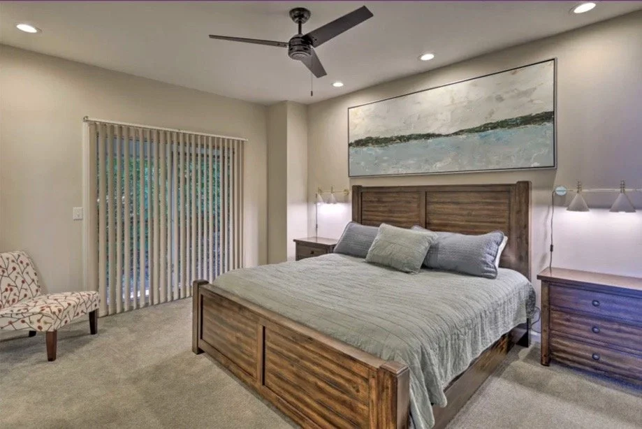 A modern bedroom with a large upholstered bed, decorative gold wall art, two bedside tables with matching lamps, a TV mounted on the wall, and a bathroom with a glass shower door visible through the open door.