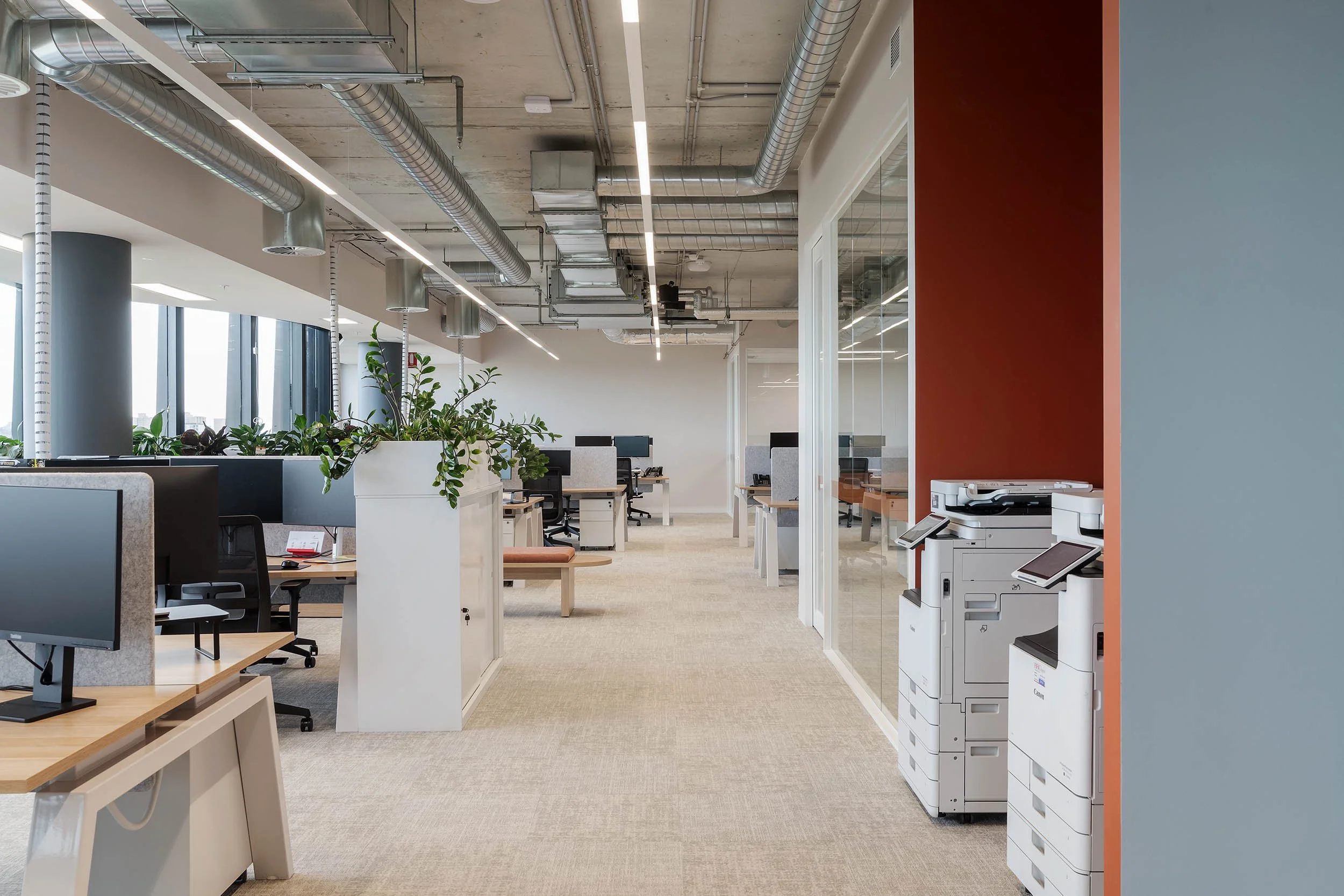 : Bright, open office layout showing rows of white workstations with dual monitors along a window line.