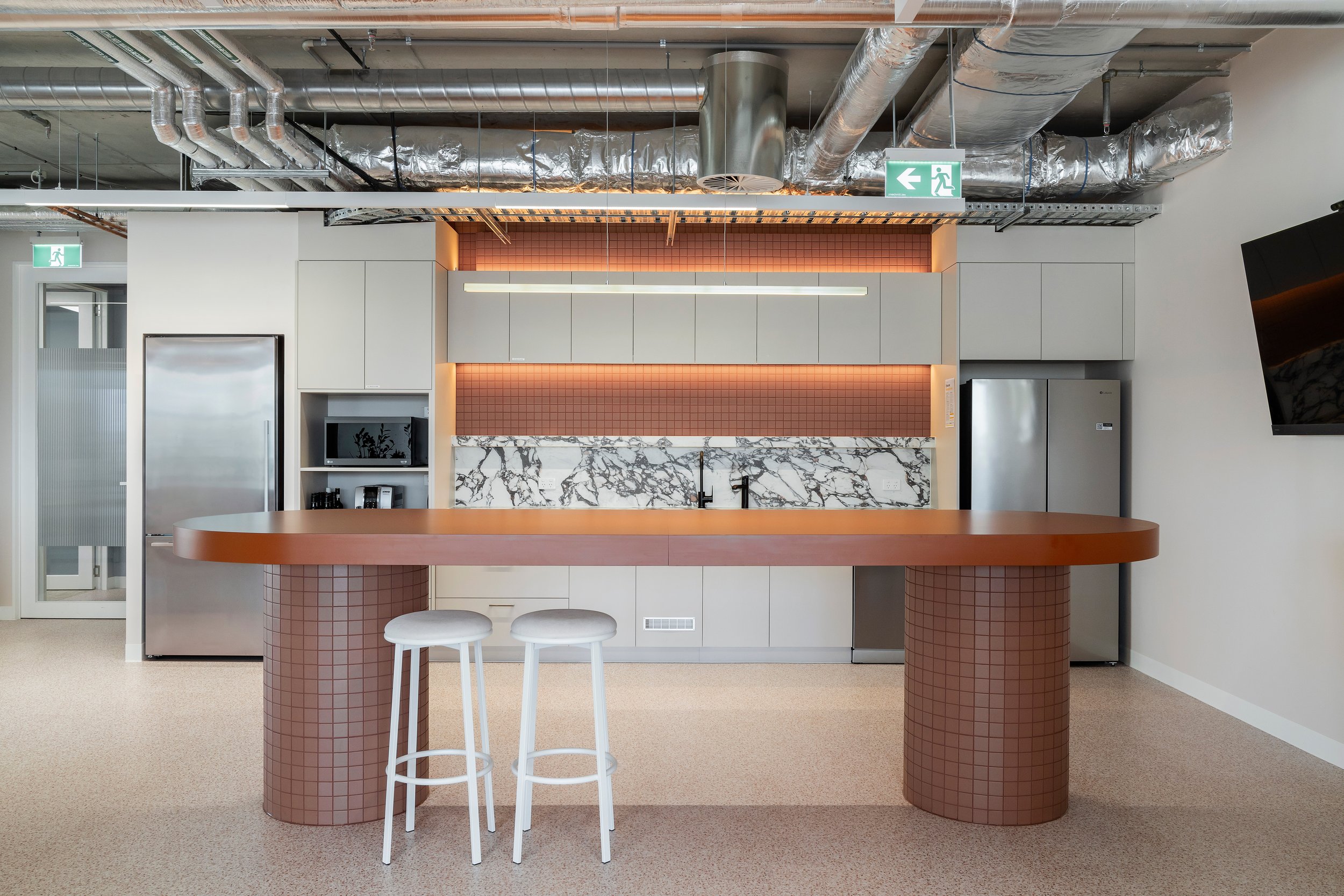 Office kitchen breakout area with a long wooden high table, white bar stools, and a marble backsplash.