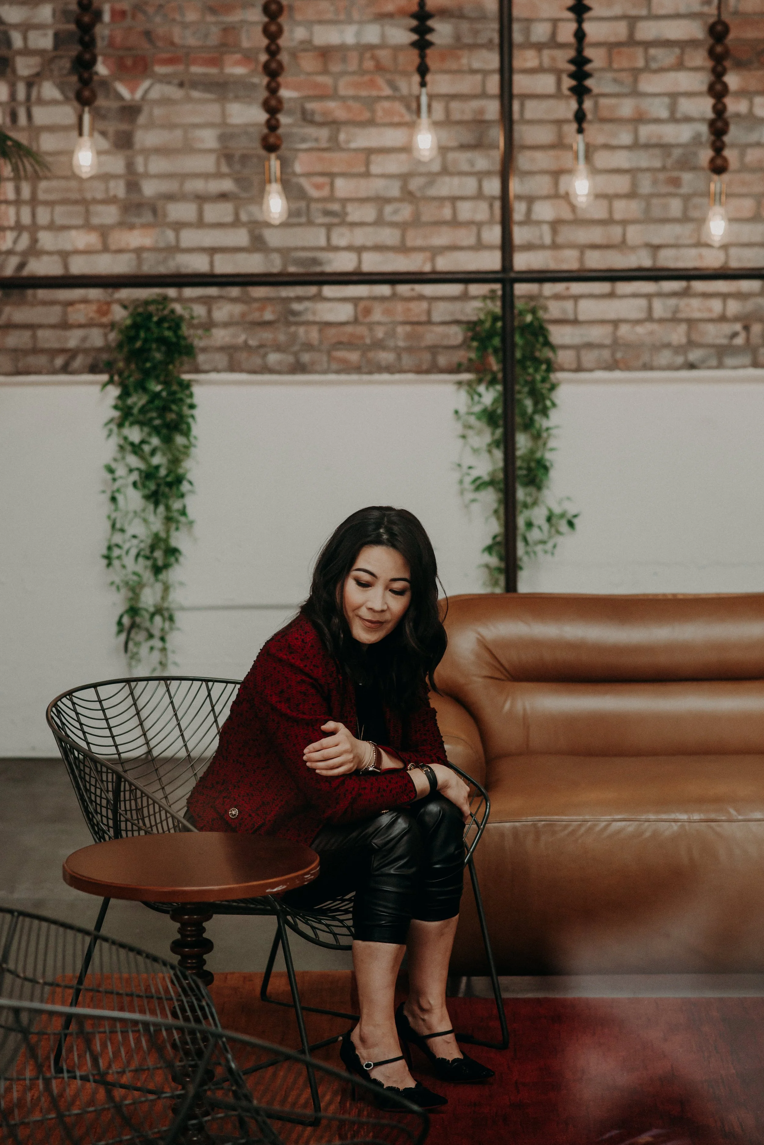 A woman with dark wavy hair sitting on a wire chair in a stylish, modern space with exposed brick wall, hanging light bulbs, and green plants. She is wearing a red blazer, black leather pants, and black heels.