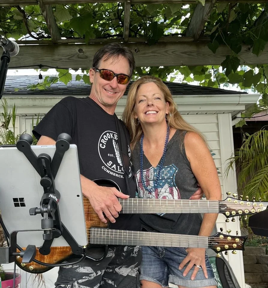 A man and woman standing together outside. The man is holding a double-neck guitar and wearing sunglasses, a black T-shirt, and patterned shorts. The woman is wearing a sleeveless top, denim shorts, and a necklace. They are smiling and posing under a wooden pergola with greenery.