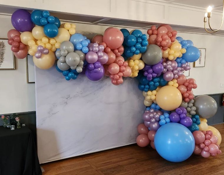 A colorful balloon arch with various-sized balloons in purple, pink, blue, yellow, and grey against a blank backdrop. The setting includes a wooden floor and framed artwork on the walls.