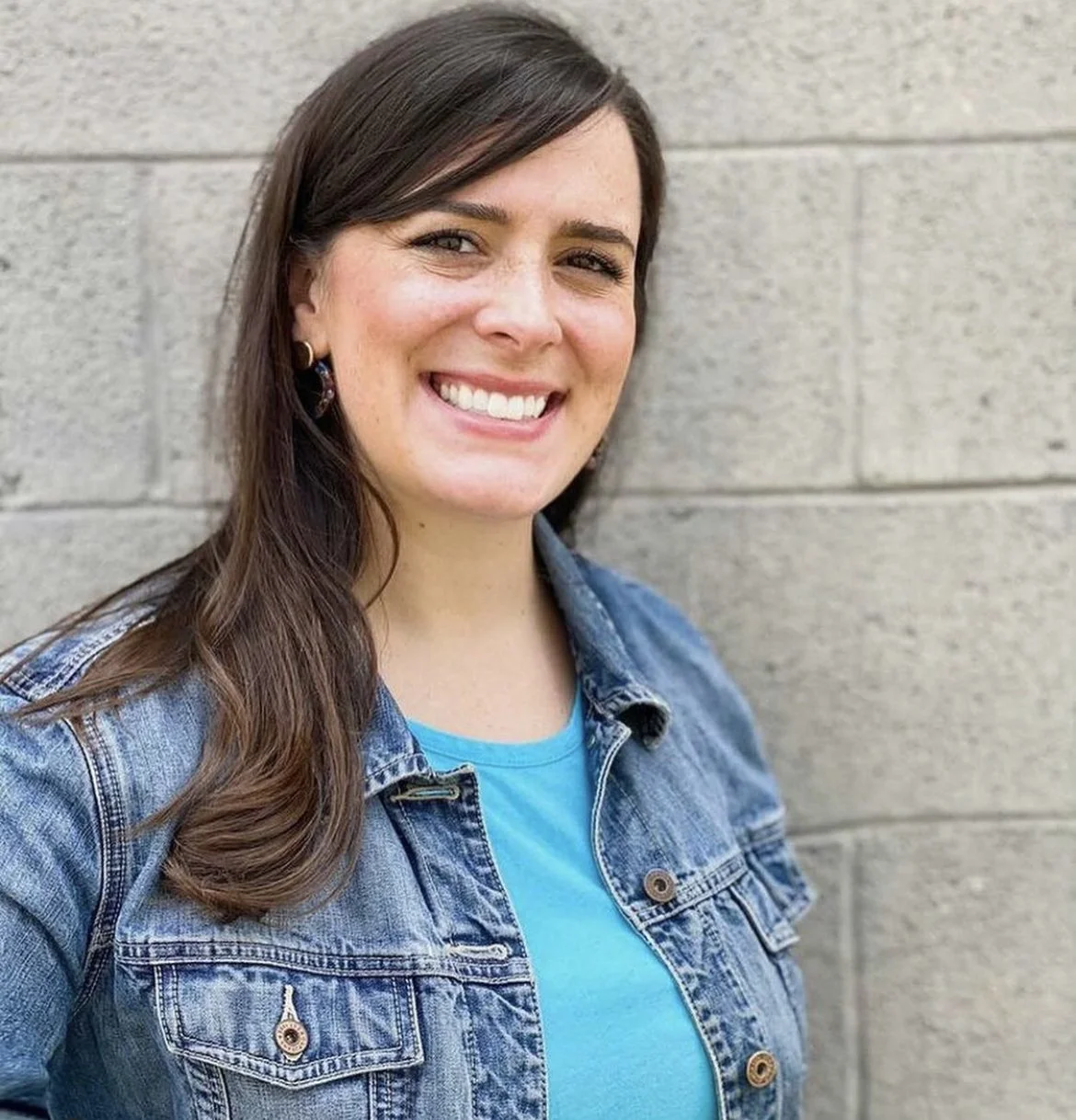 Smiling person wearing a blue shirt and denim jacket standing in front of a gray brick wall.