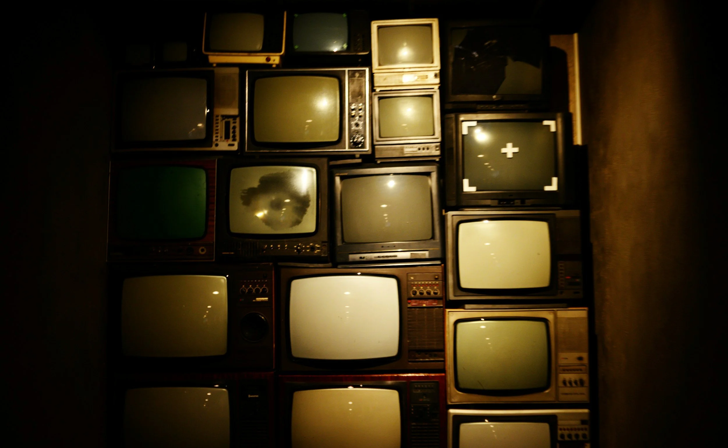 A wall of vintage CRT televisions and monitors stacked together in rows, showcasing various sizes and styles in a dimly lit setting.