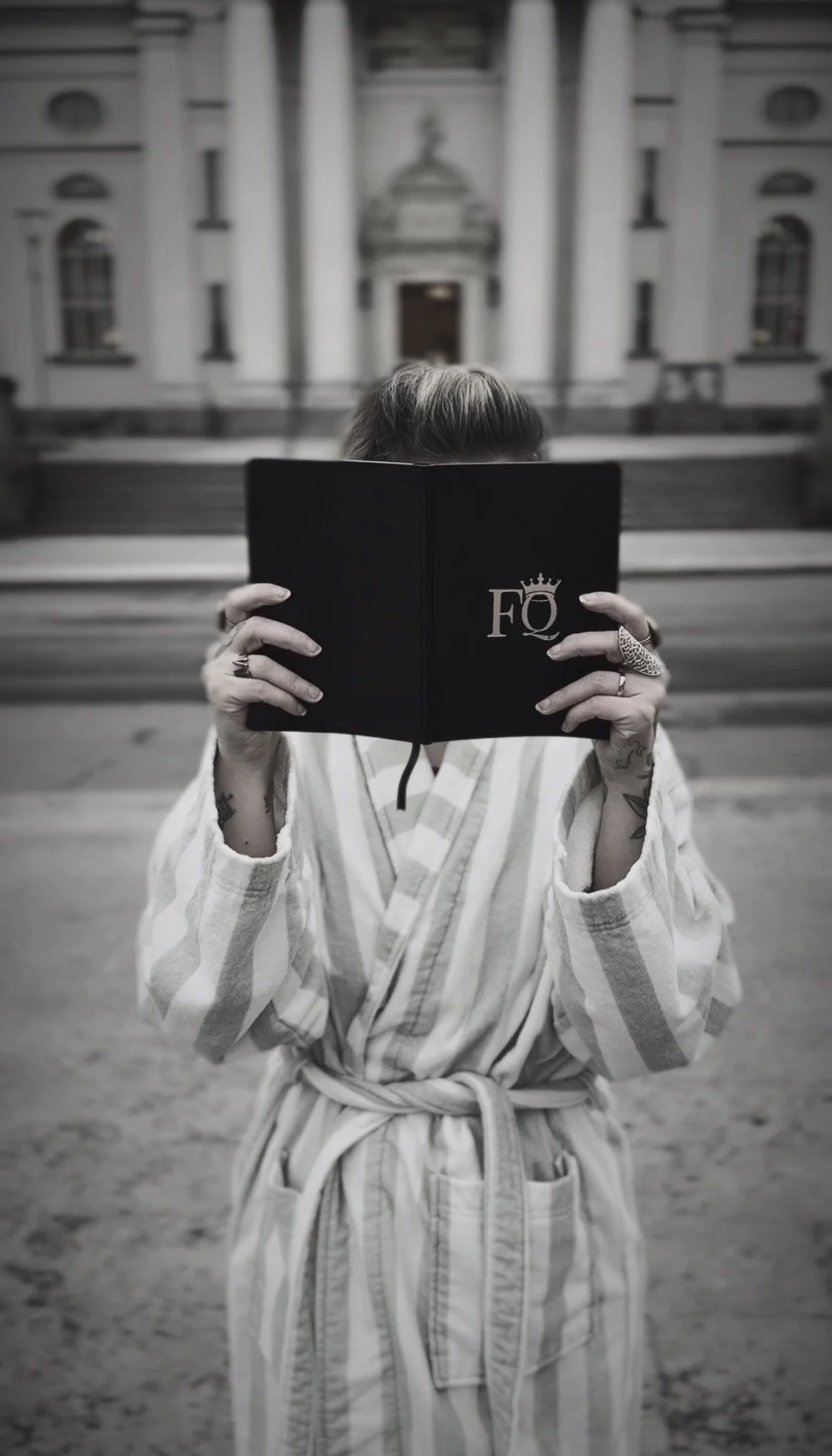A person wearing a striped robe and various rings on their fingers is holding a black book with the letters 'FO' and a crown symbol on the cover, covering their face. The background features a historic building with columns.
