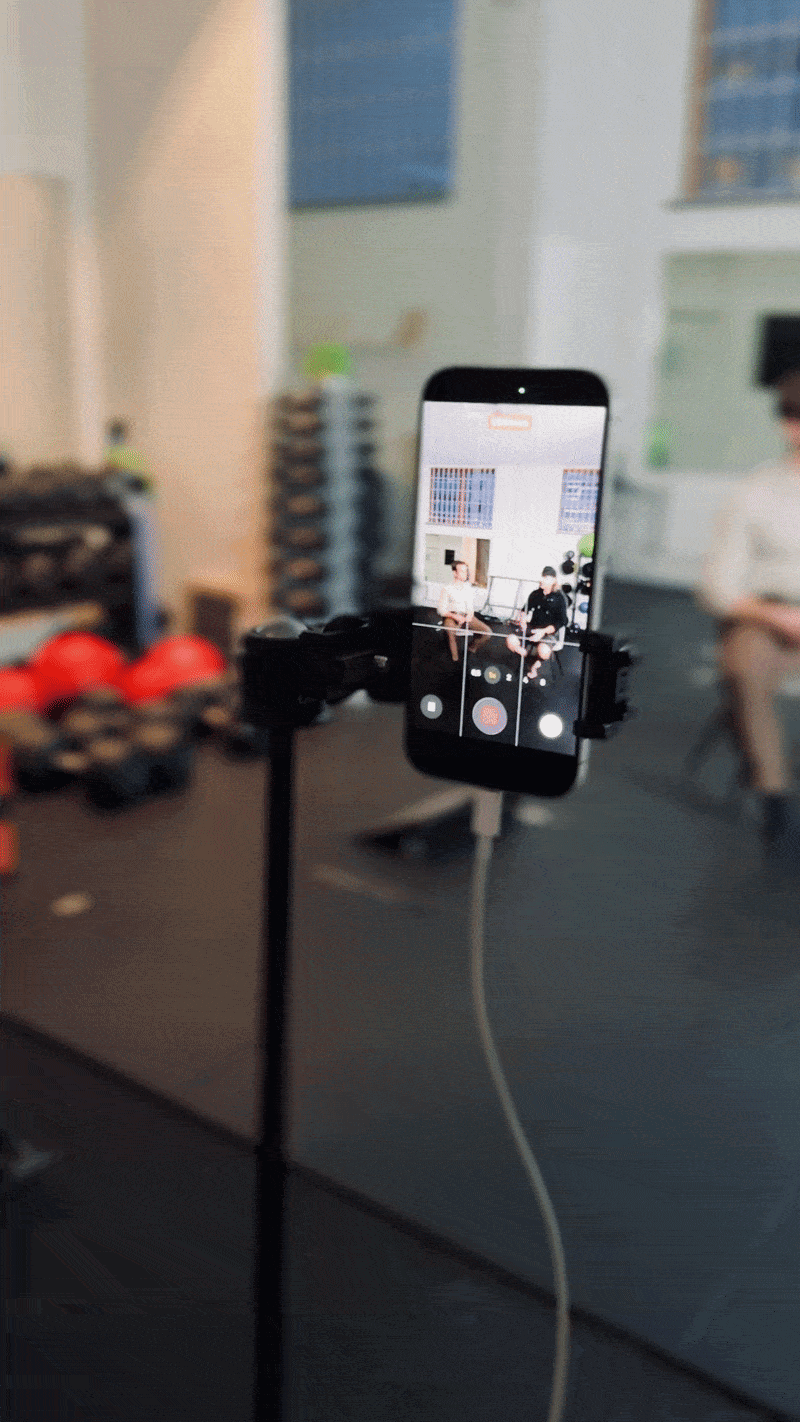 Smartphone mounted on a stand recording two people sitting on chairs in a room with posters on the wall.
