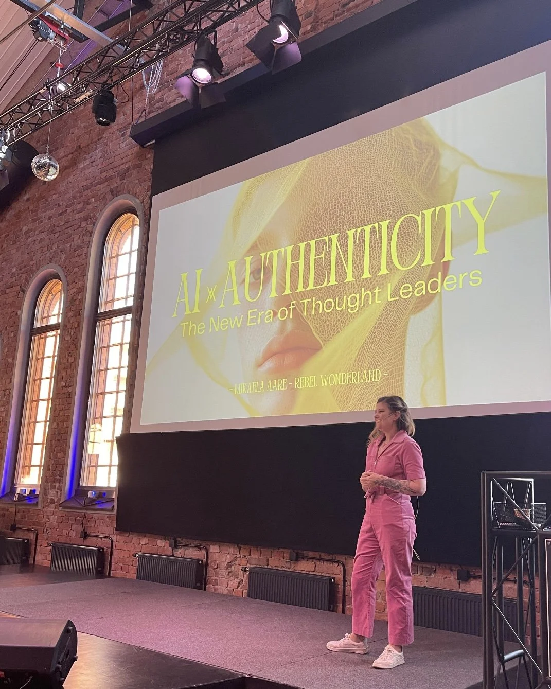 A woman stands on stage in front of a large presentation screen with the title 'AI AUTHENTICITY: The New Era of Thought Leaders' by Mikaila Aare at Rebel Wonderland. The stage is in a brick-walled venue with arched windows and stage lighting.