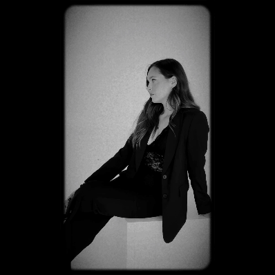 A woman sitting on a ledge against a plain background, wearing a black blazer and pants, facing to the side in black-and-white.