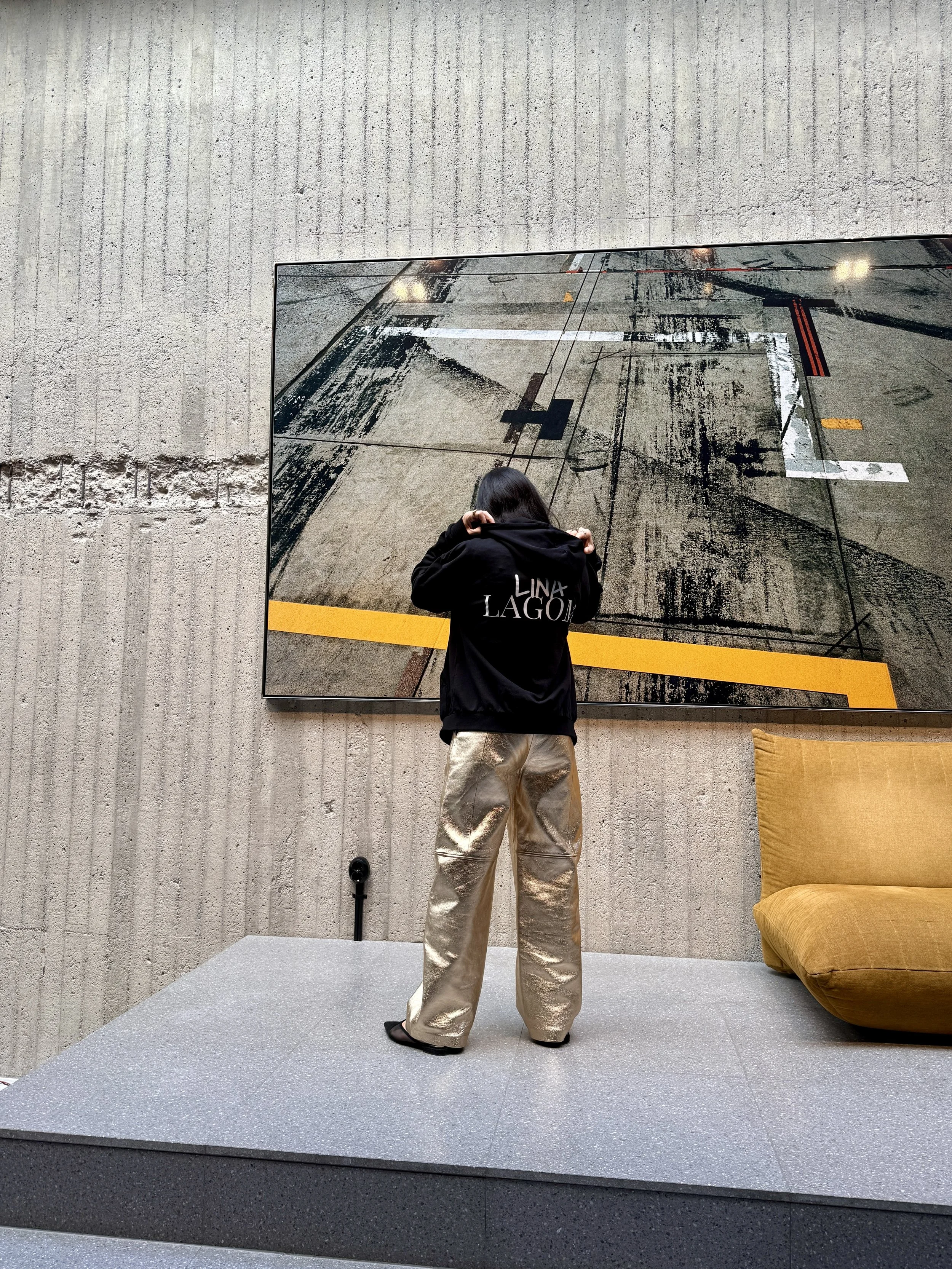 A person with long dark hair wearing a black hoodie with the words "LINAX LAGO" printed on the back, and shiny gold pants, standing on a raised platform in front of a large mirror showing their reflection, against a gray textured wall, with a mustard yellow cushion on a chair to the right.