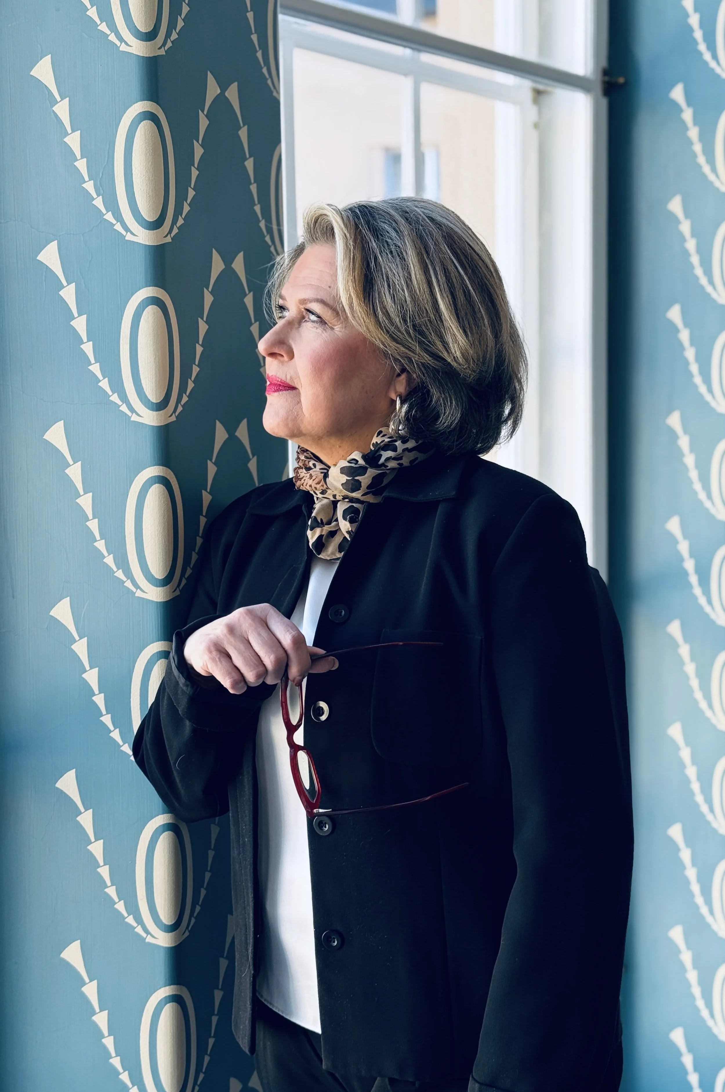 A woman with shoulder-length gray hair, wearing a black blazer and a leopard print scarf, stands by a window with sunlight shining on her face, holding red glasses in her hand, with a blue patterned wall behind her.