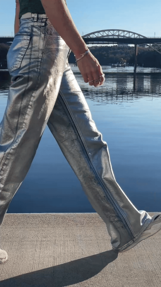 Close-up of a person walking by a body of water with a bridge in the background, wearing shiny silver pants and a black top.