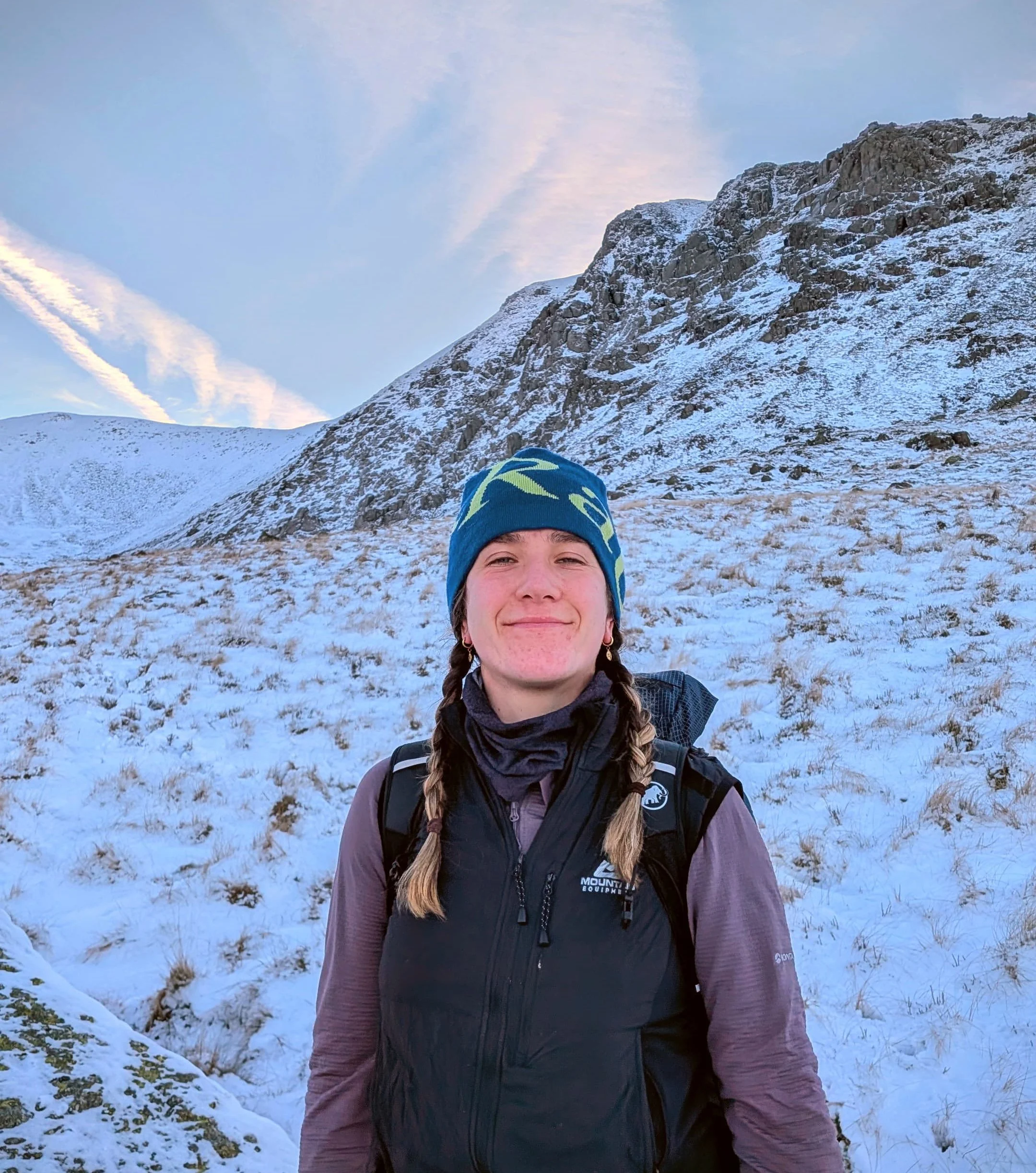 A woman with braided hair smiling in a snowy mountain landscape while wearing a blue beanie, a black outdoor vest, and long-sleeved shirt.