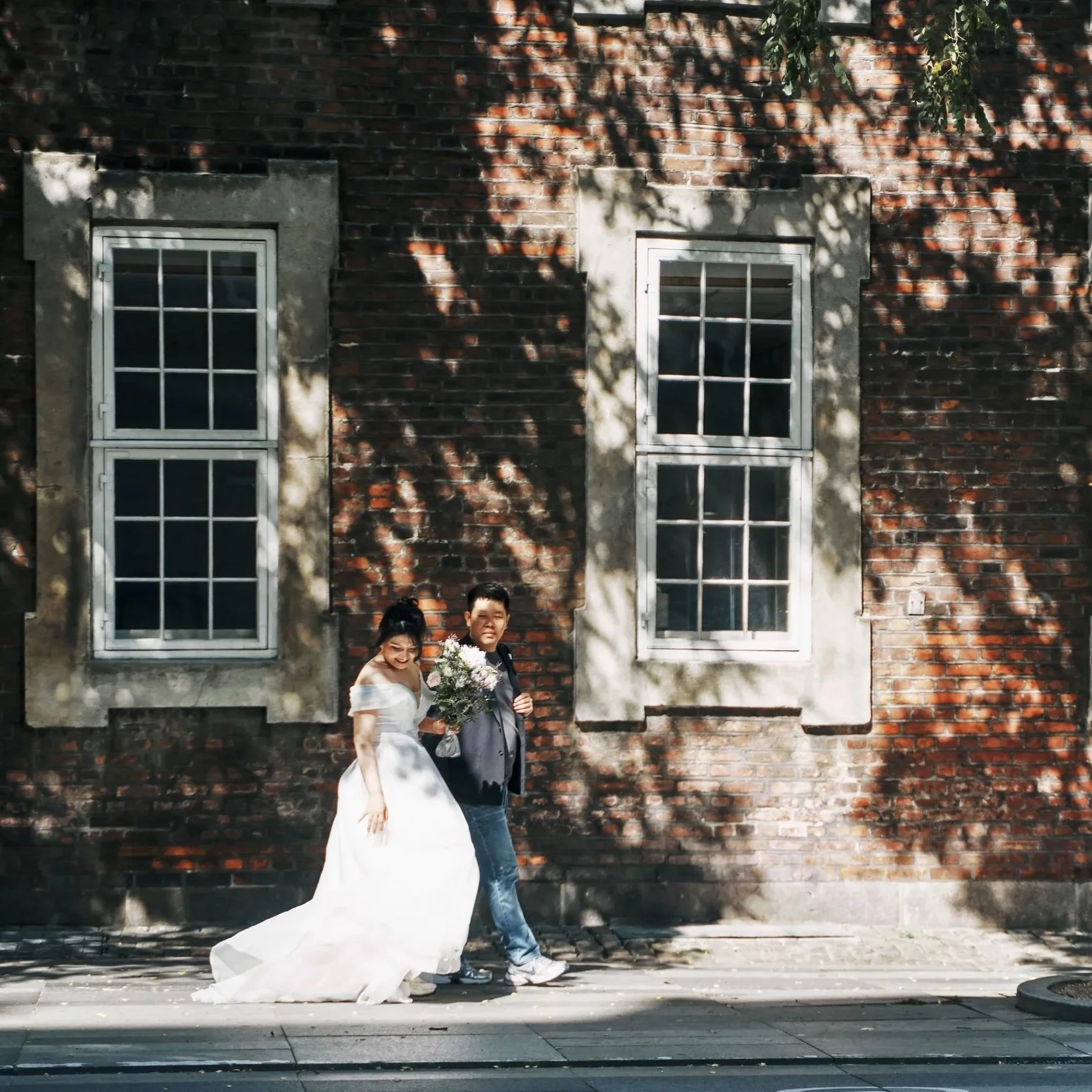 A man and woman sitting on stairs indoors. The woman wears a traditional Chinese Hanfu, featuring a red, blue, and gold floral pattern. The man wears dark casual clothing. Both are smiling, with a bright skylight above.