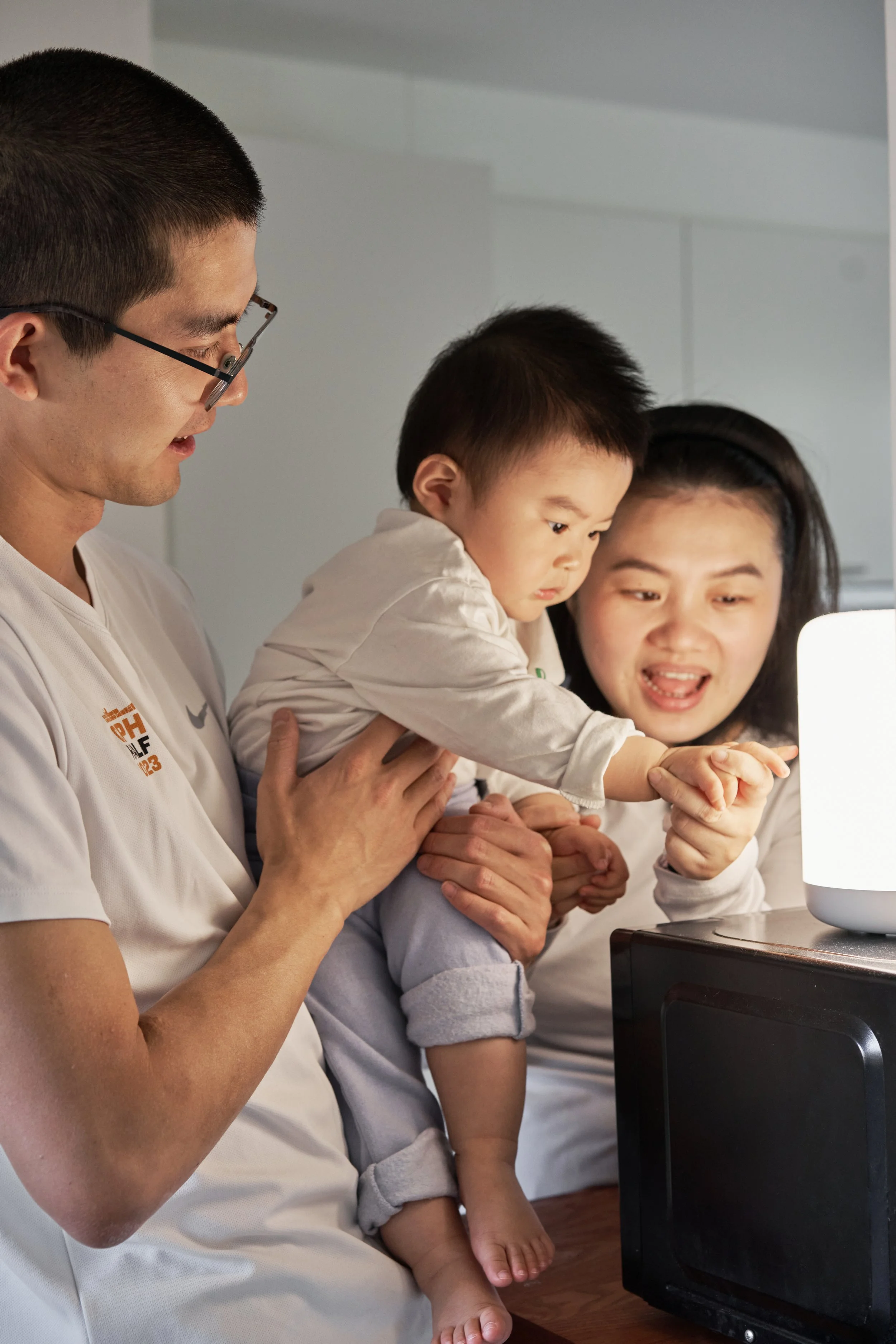 A family in a kitchen with a father holding a young child who is touching a lamp, while the mother smiles and guides the child's hand.