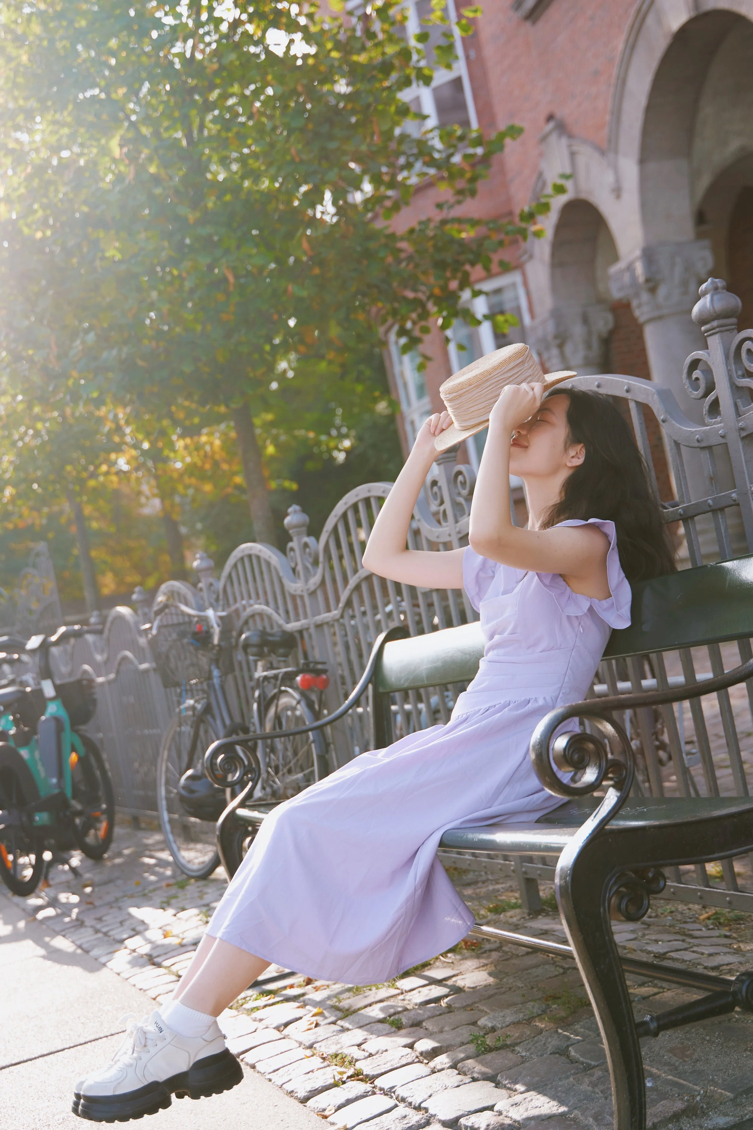 Woman in a light purple dress sitting on a bench, holding a straw hat, with sunlight filtering through trees, bicycles and a building in the background.