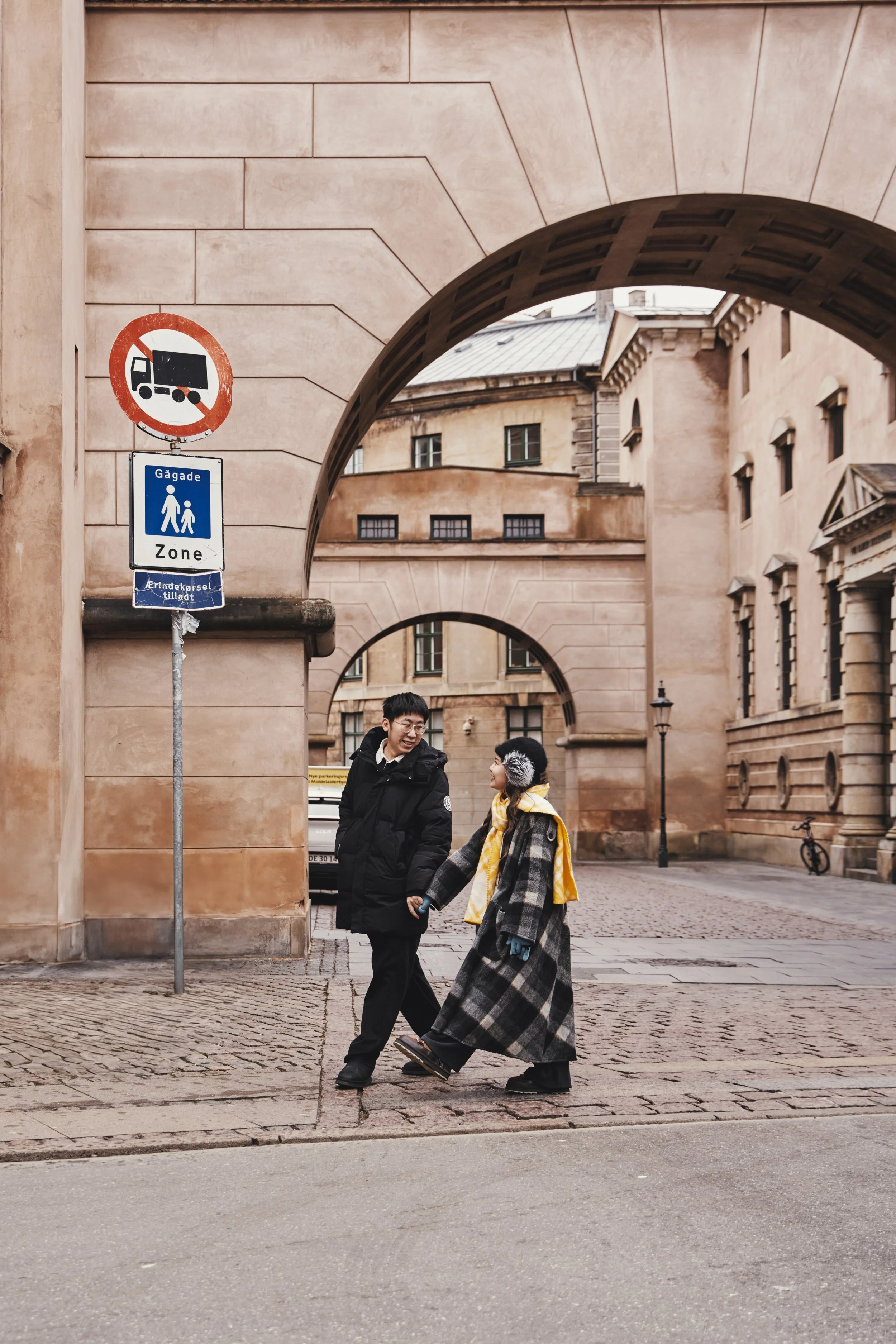 Woman in a light purple dress sitting on a bench, holding a straw hat, with sunlight filtering through trees, bicycles and a building in the background.