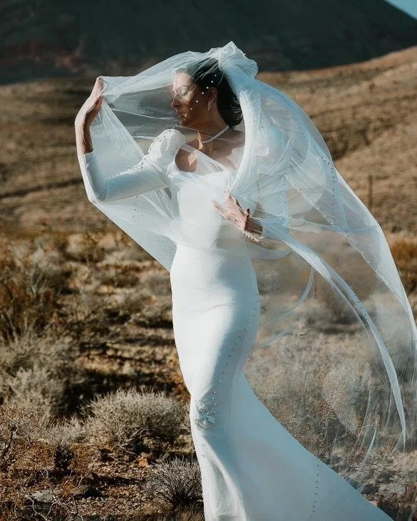 Woman in a white wedding dress with long sleeves and a veil, standing in a desert landscape with mountains in the background.