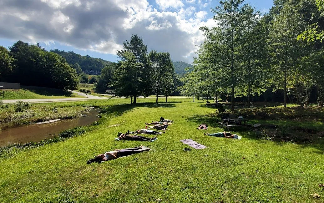 People practicing yoga in a grassy park near a small stream, surrounded by trees, with mountains and partly cloudy sky in the background.