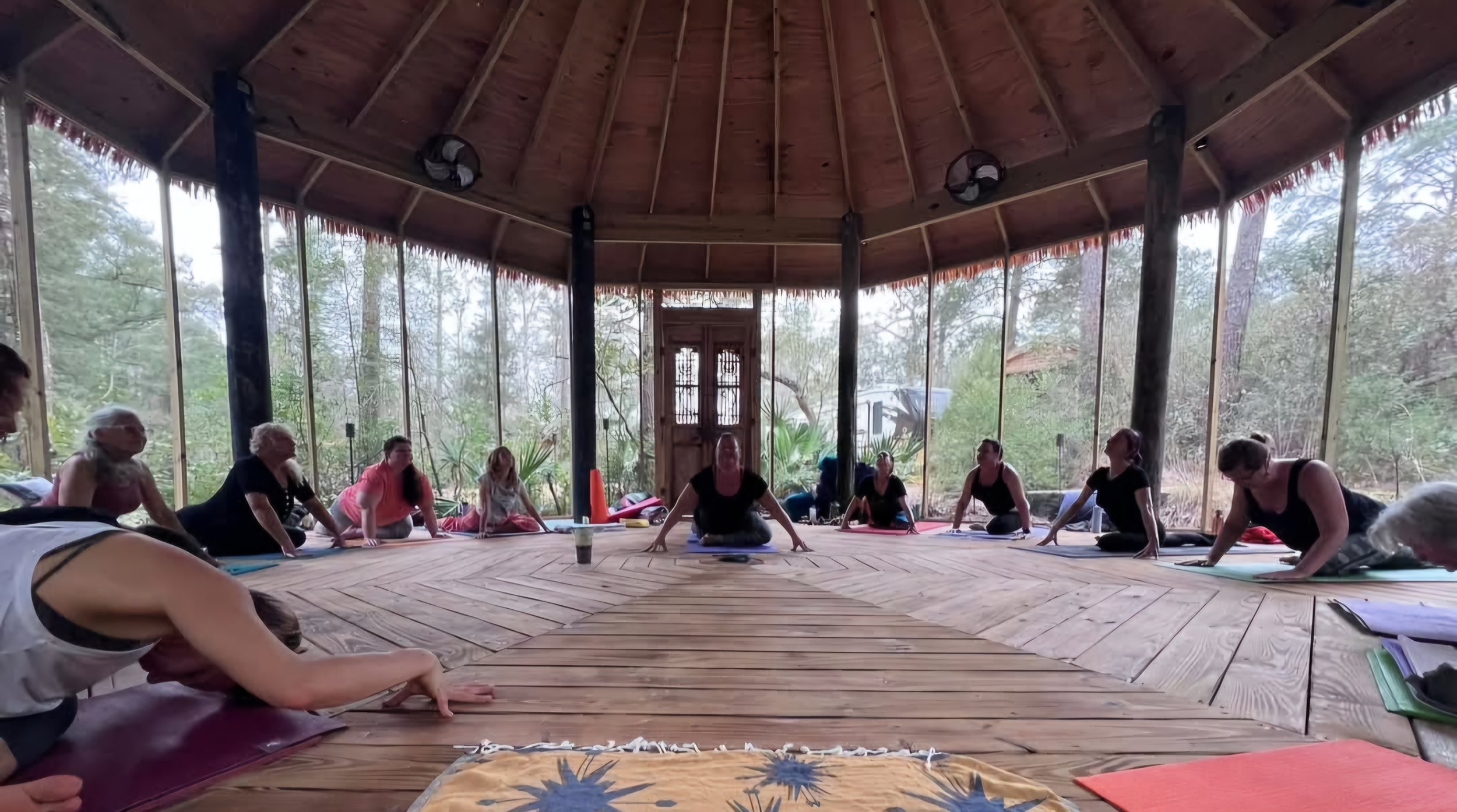 Group of people participating in a yoga or meditation class inside a circular, open-air wooden structure surrounded by trees.