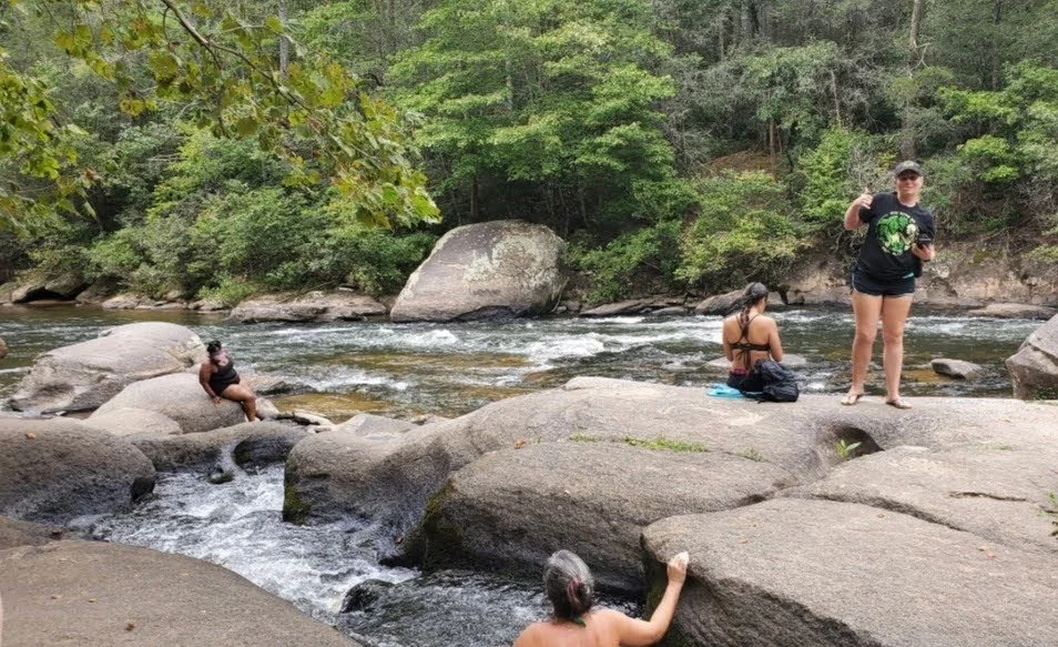 Four people relaxing by a river with large rocks and lush green trees in the background.