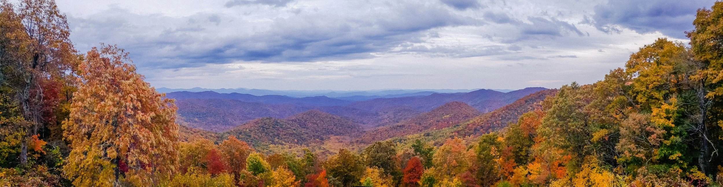 A scenic view of a mountain range covered in autumn foliage with trees in orange, yellow, and red in the foreground and layered mountains in the background under a cloudy sky.