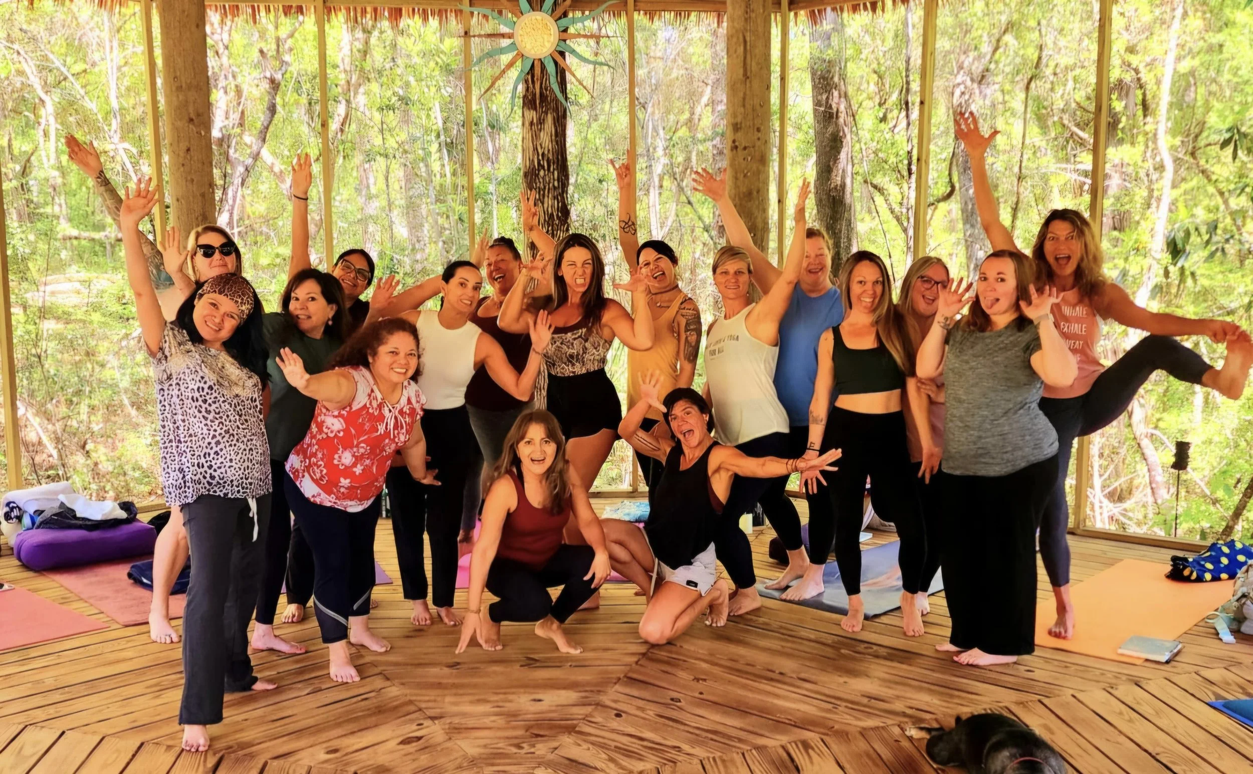 Group of smiling women and men posing together in a wooden room with large windows showing trees outside, some making playful gestures and one person lifting another's leg.