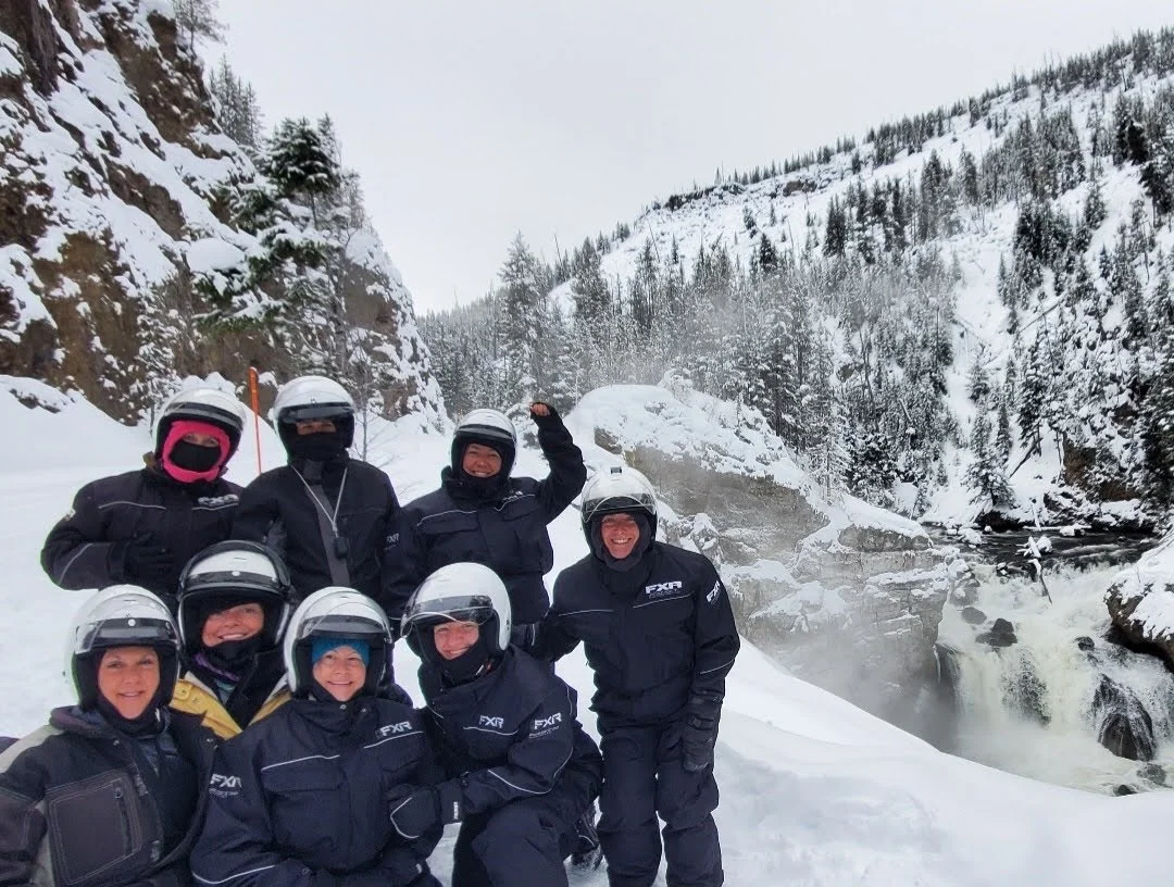 A group of eight people in winter gear and helmets posing in front of a snowy landscape with a river, rocks, and snow-covered trees and mountains in the background.