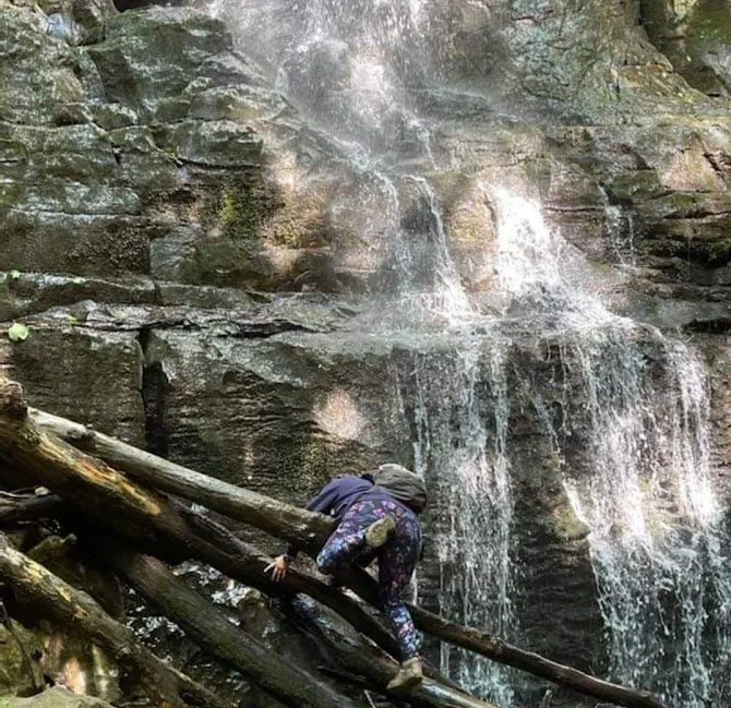A person wearing dark clothing and a cap is climbing on logs placed against a rocky waterfall wall.