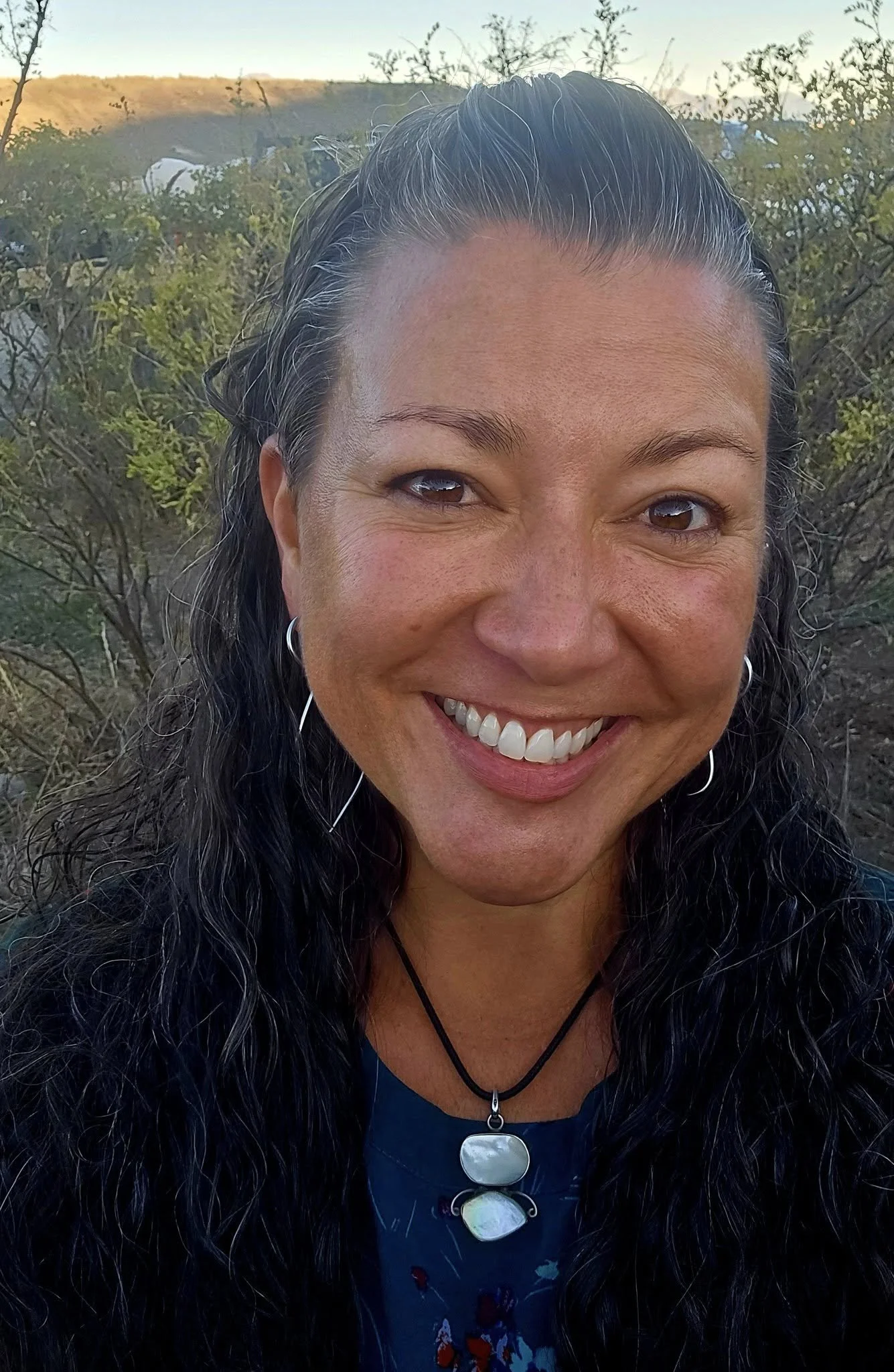 Close-up of a smiling woman with long, wavy black hair wearing a necklace with three pendant stones, outdoors with a background of bushes and distant hills.