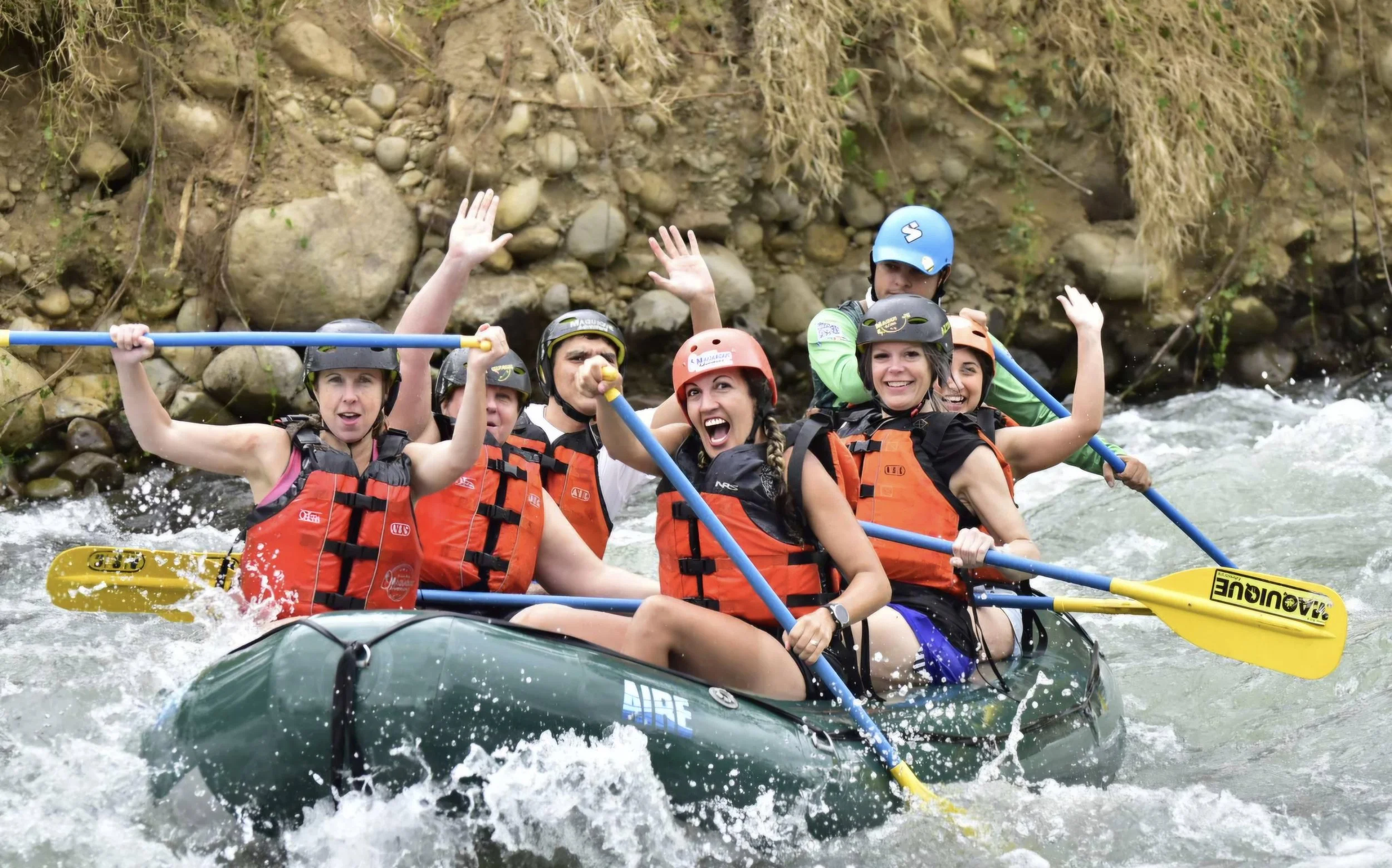 Group of people white water rafting on a river, wearing life jackets and helmets, with rocky riverbank in background.